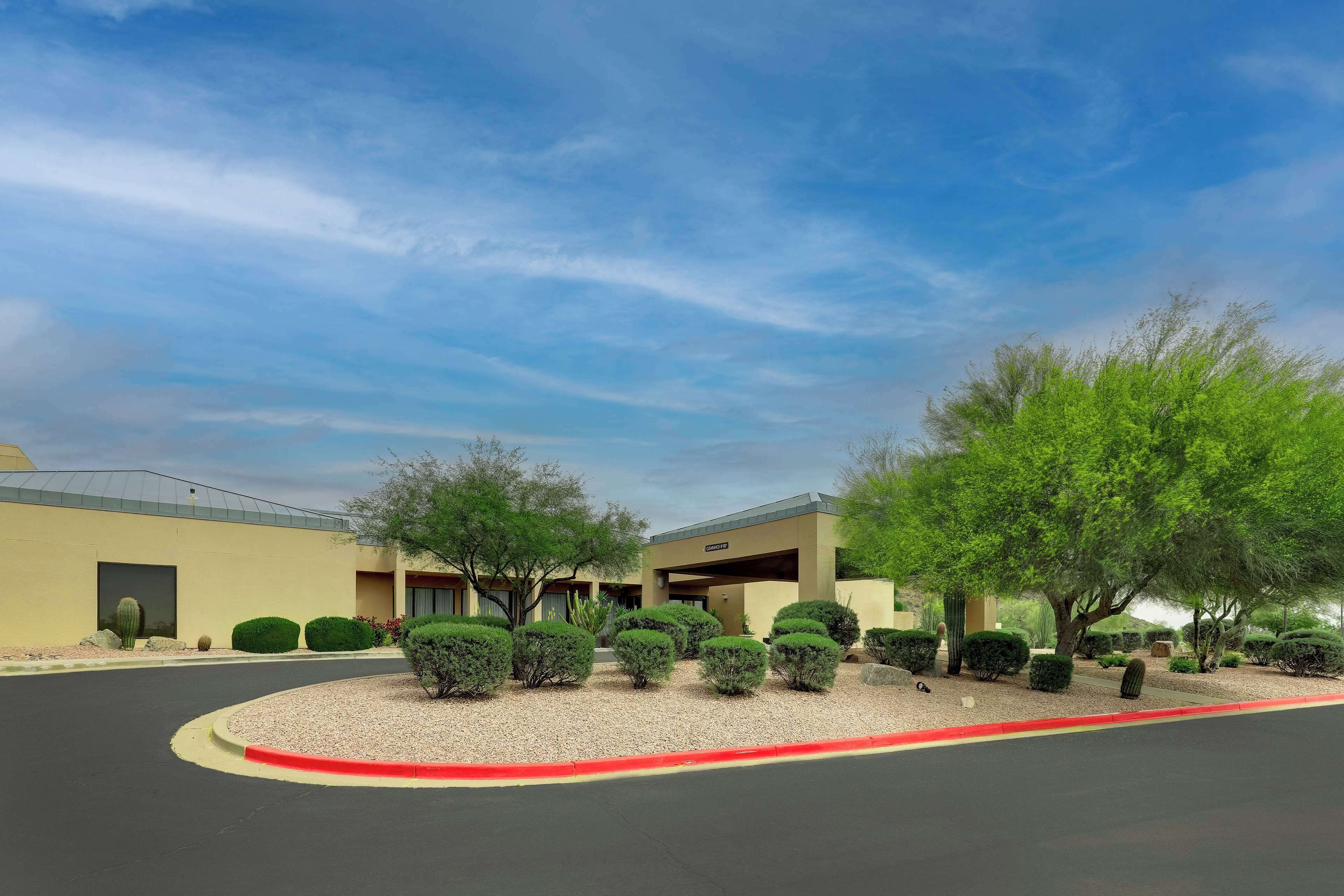 A building with a modern design, featuring a curved driveway and landscaped areas with various shrubs and trees.