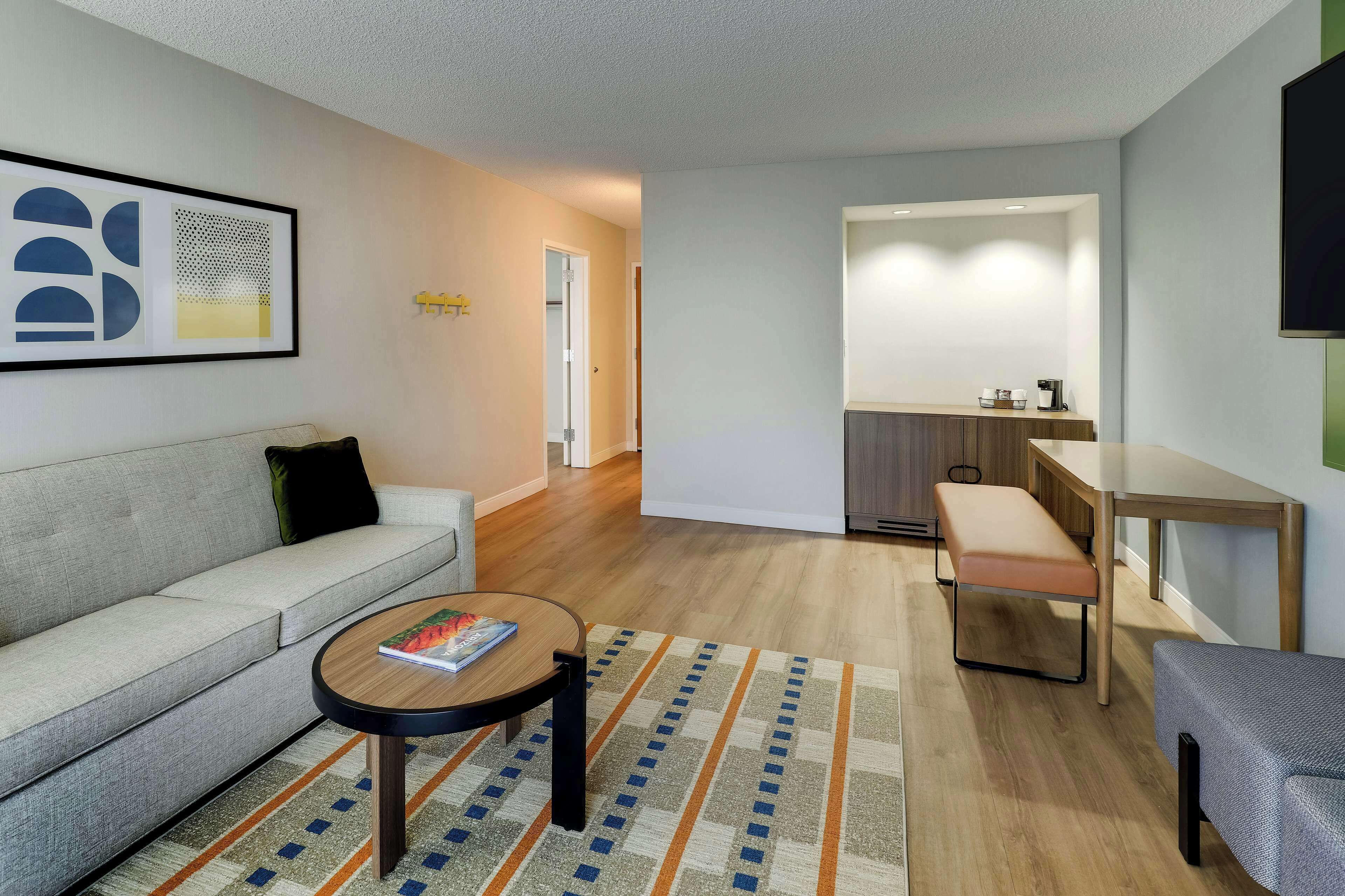A modern living room with a sofa, coffee table, and a rug. There is a kitchenette area with a bench and a TV mounted on the wall.