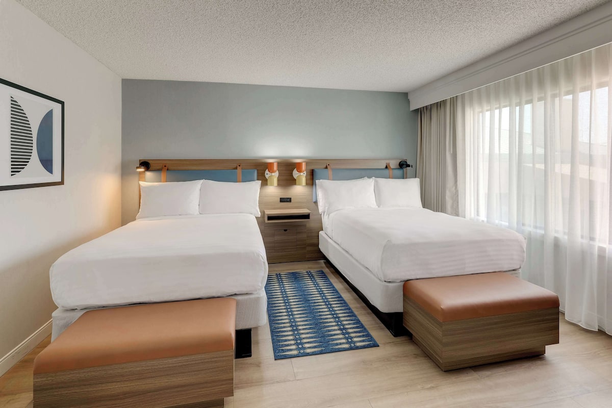 Een hotelkamer met twee bedden, een nachtkastje, een blauw vloerkleed en een raam met dunne gordijnen.