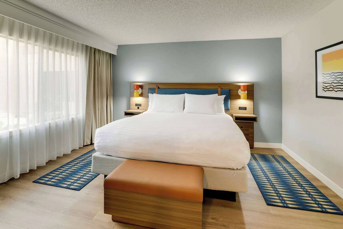 Een hotelkamer met een groot bed, bedlampen, een houten bankje en een kamerkleed met een patroon.