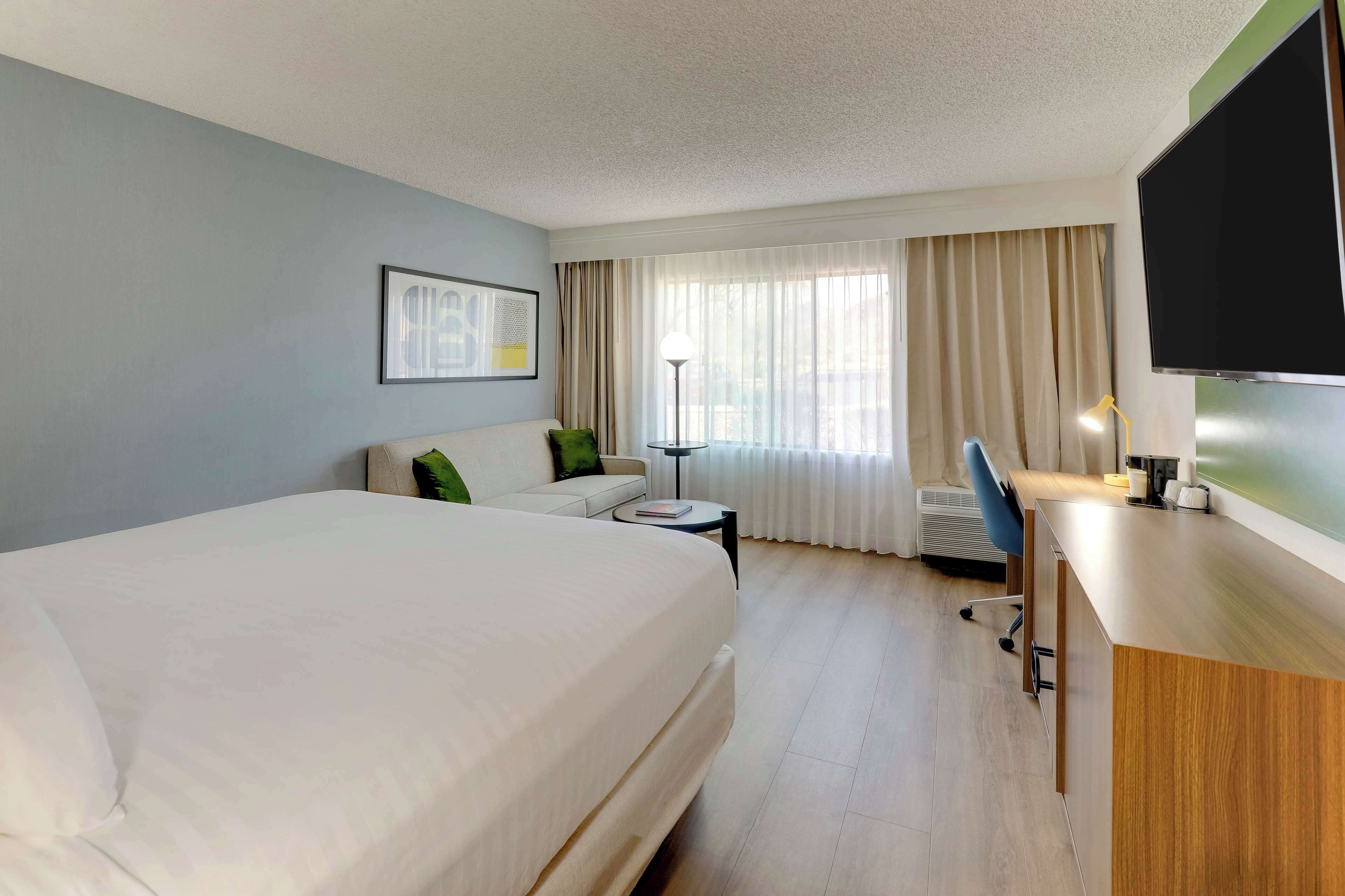 A hotel room with a large bed, a desk, a chair, a small table, a lamp, and a TV.