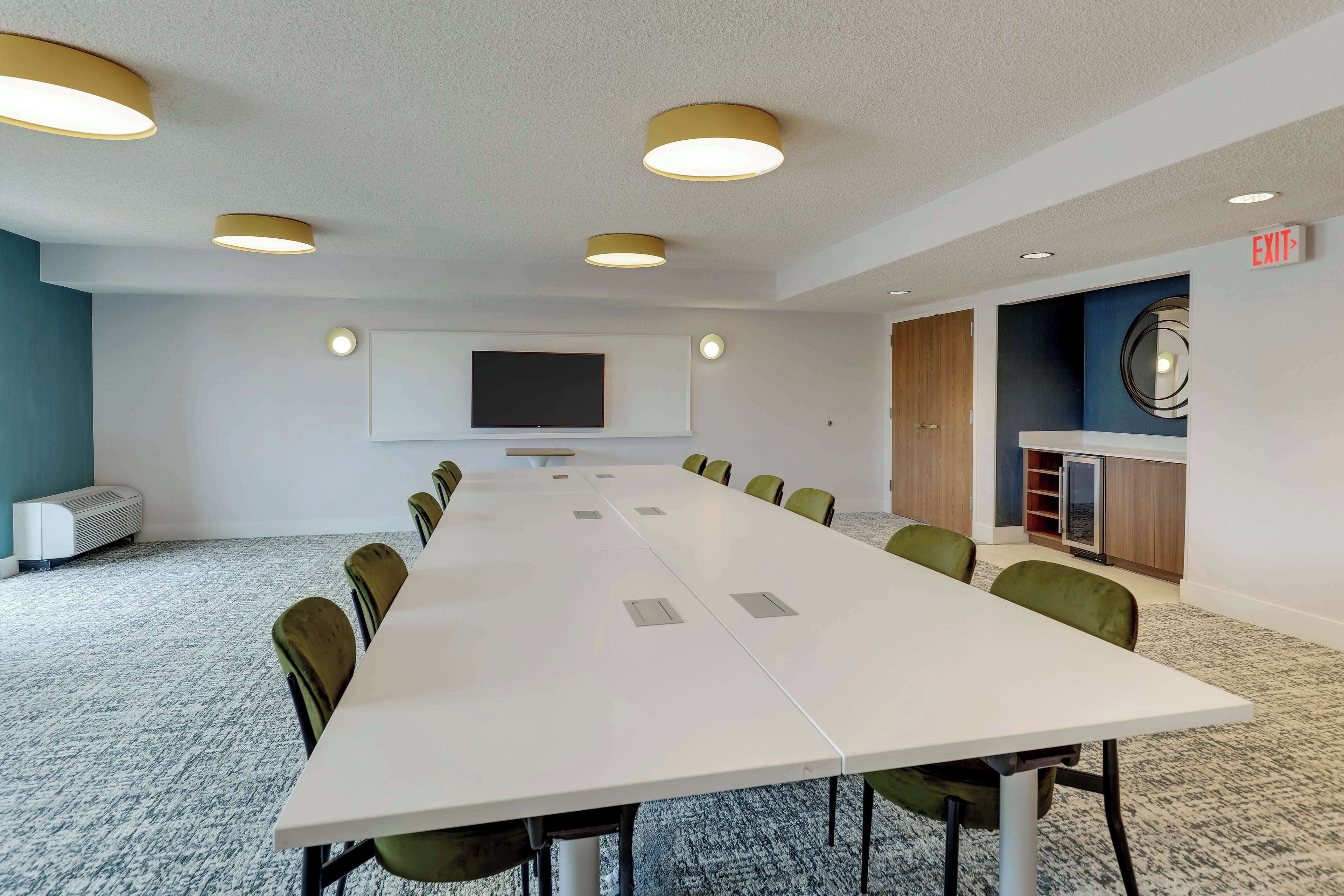 Una sala de conferencias con una mesa larga, sillas verdes, un televisor de pantalla plana y una pequeña área de cocina.