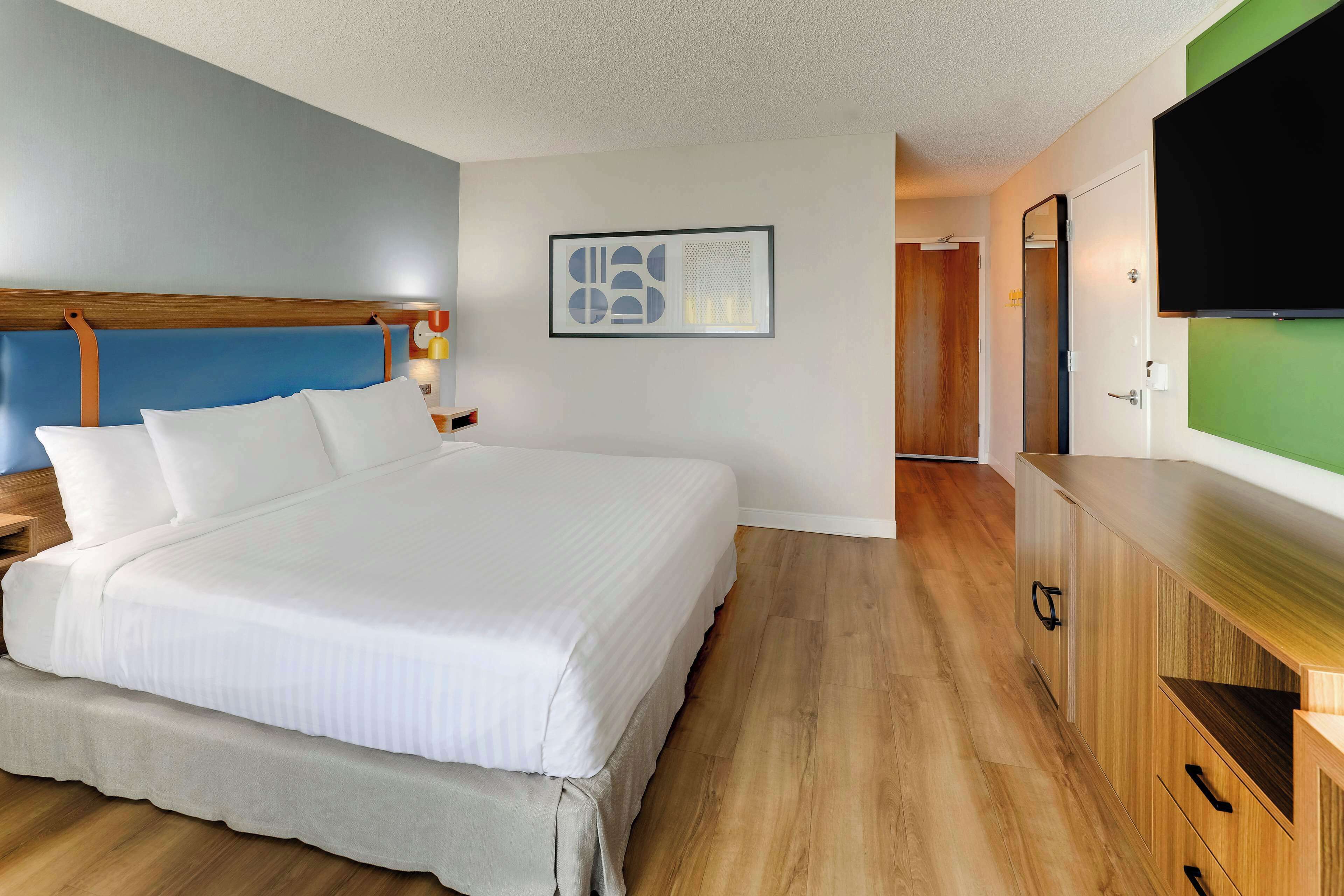 A hotel room with a large bed, wooden headboard, a flat-screen TV, a wooden dresser, and a framed abstract artwork on the wall.