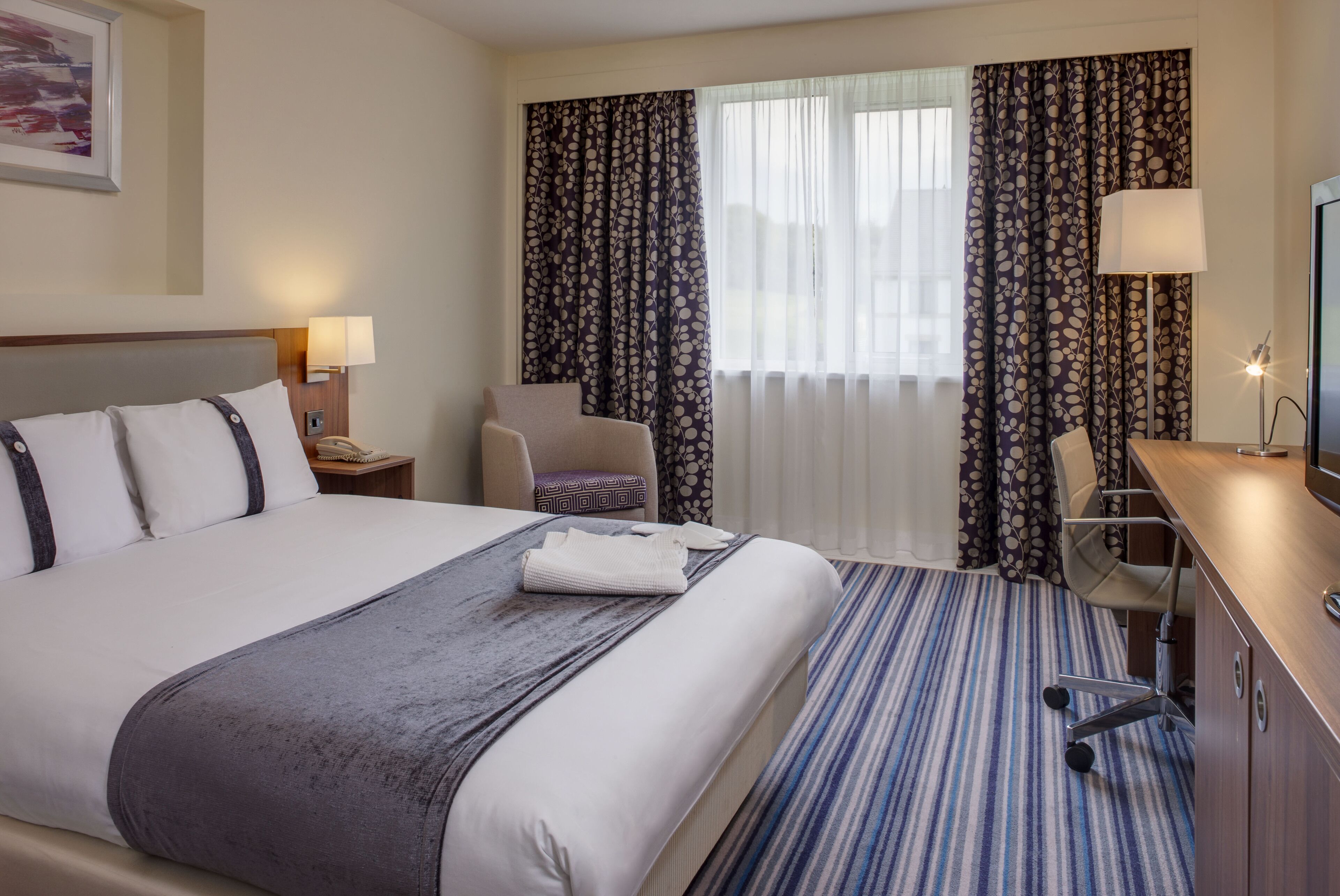 A hotel room with a large bed, a desk, a chair, and a TV.