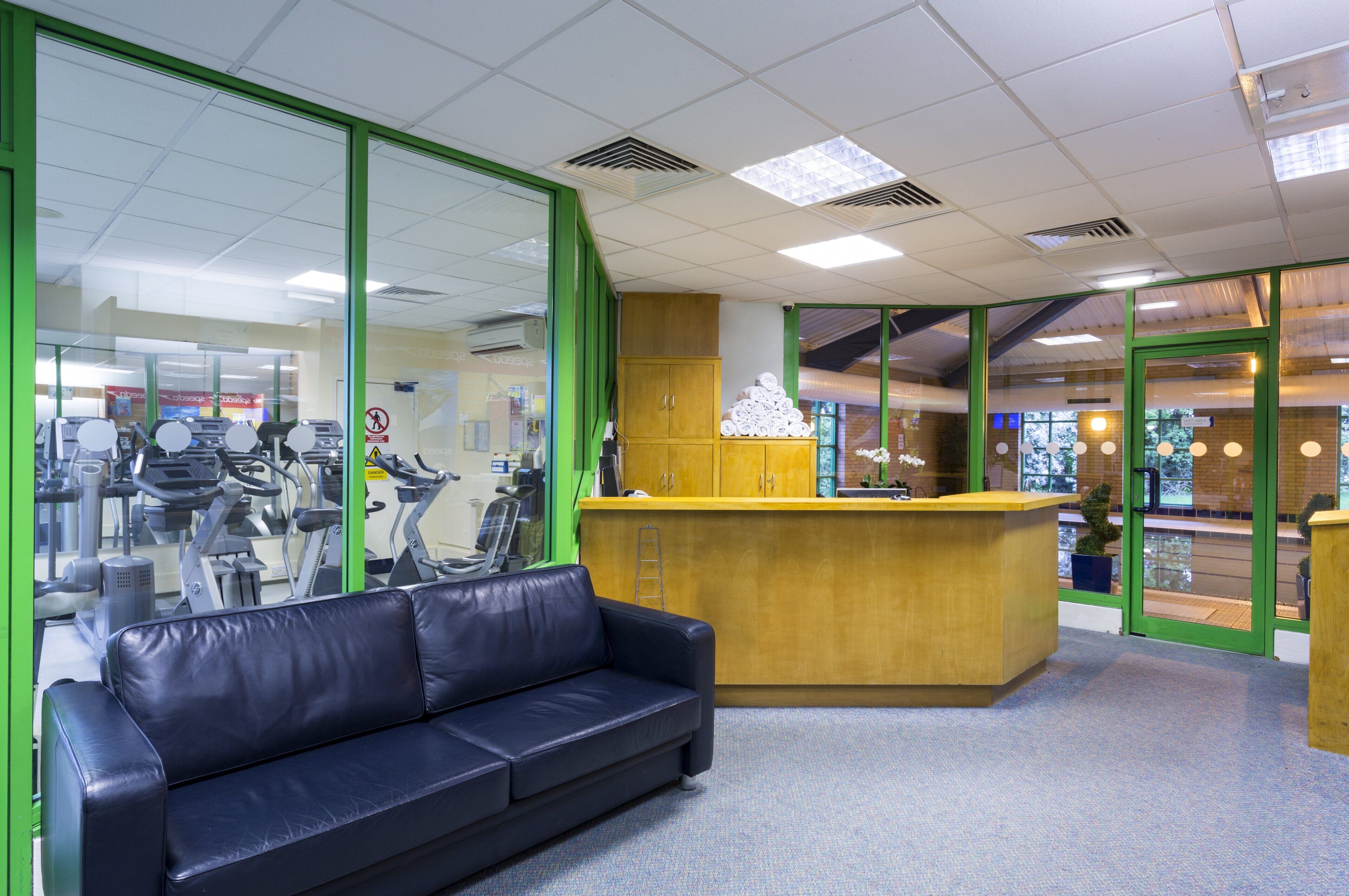 A reception area with a reception desk, a black leather couch, and a glass wall reflecting gym equipment.