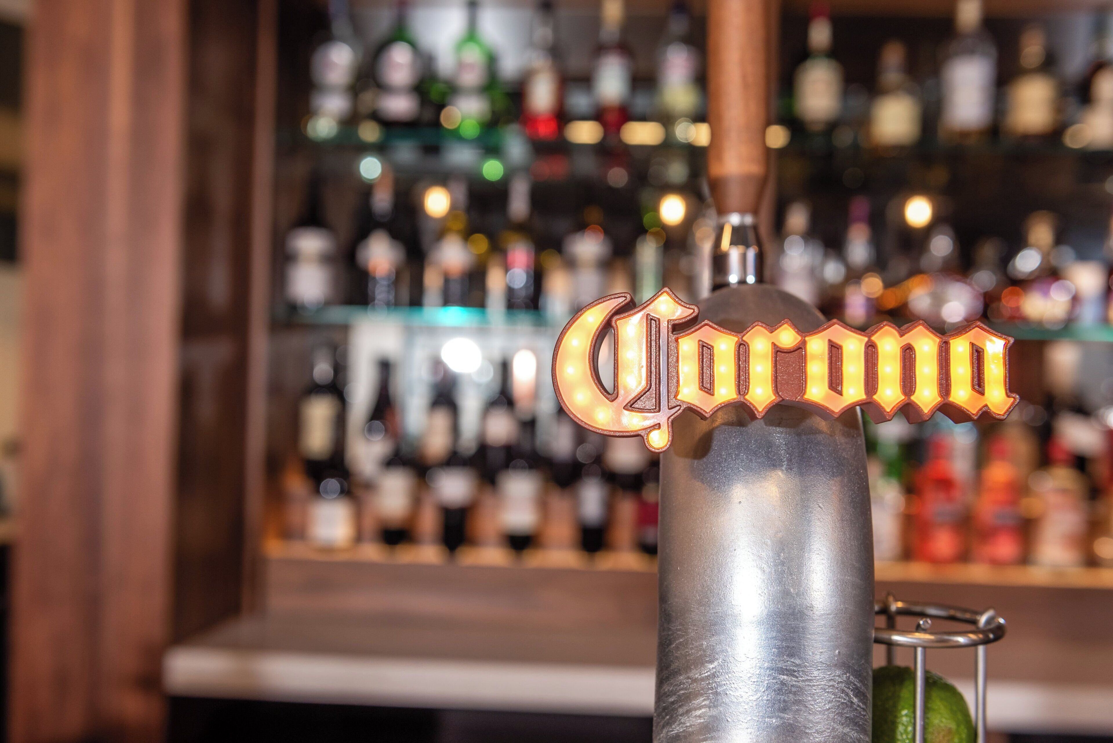 A bar setting with a prominently displayed illuminated 'Corona' sign on a metal column.