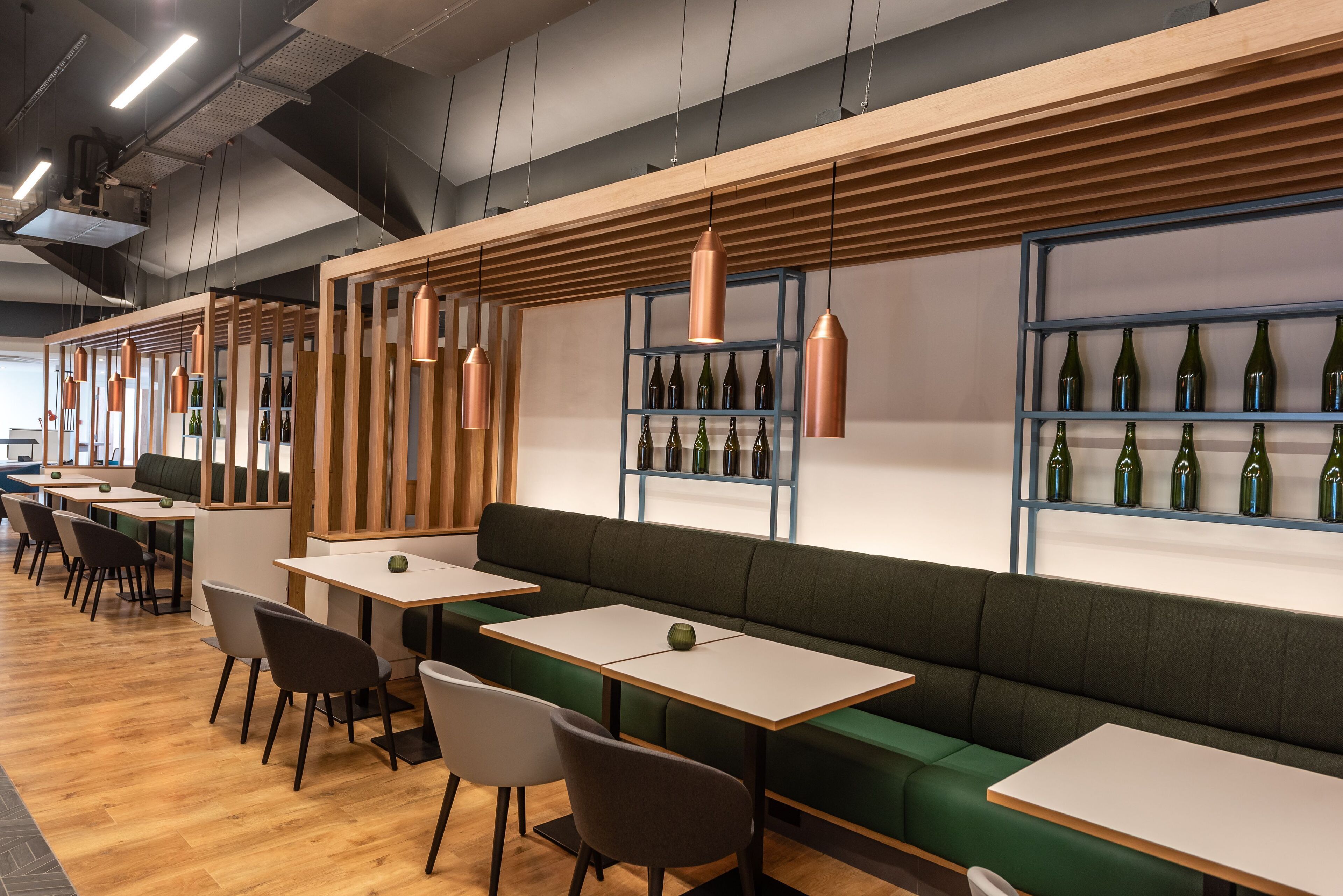 A modern restaurant with wooden slats, green cushioned seating, and copper pendant lighting. Tables are arranged with chairs.
