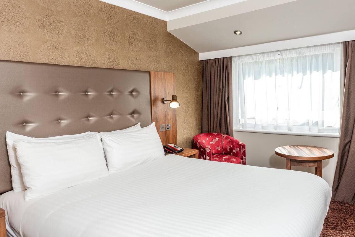 A hotel room with a large bed, a wooden headboard, a red chair, a small wooden table, and a window with curtains.