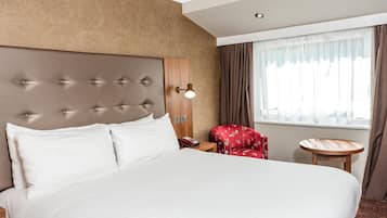A hotel room with a large bed, a wooden headboard, a red chair, a small wooden table, and a window with curtains.