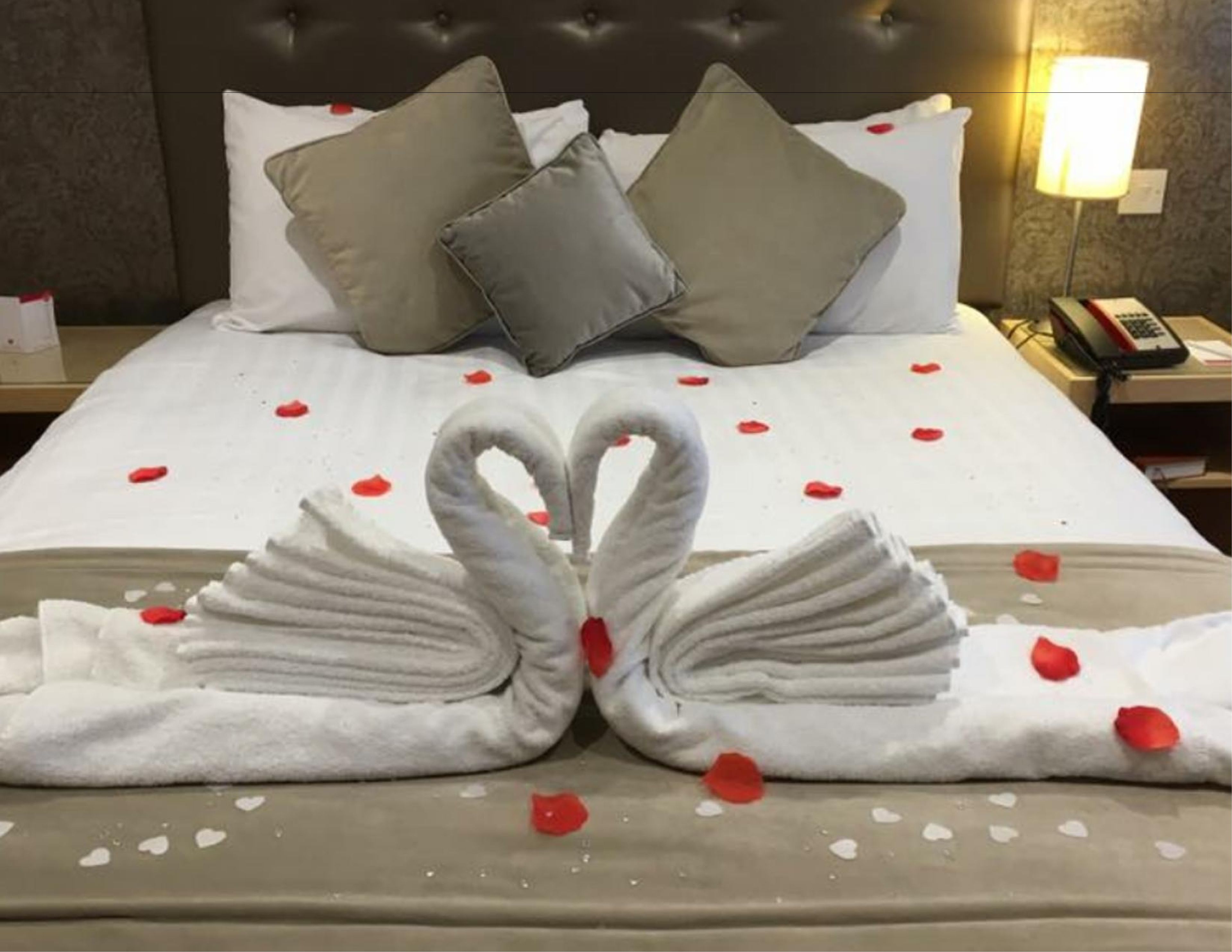 A neatly made bed with a swan towel arrangement and rose petals on a hotel room.