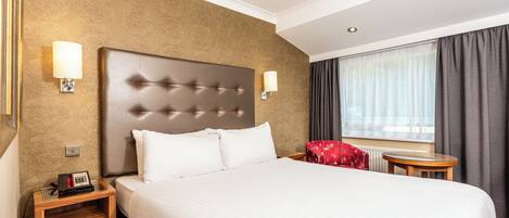 A hotel room with a large bed, two bedside tables, a red chair, a window with curtains, and a textured wall.