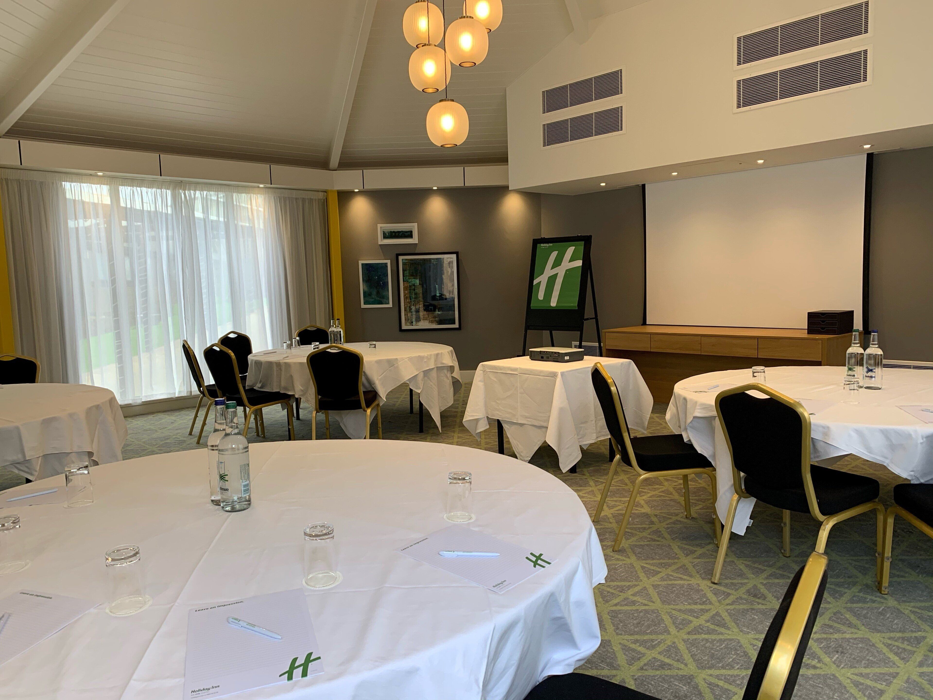 A hotel conference room with round tables, chairs, a projector screen, and a large window with curtains.