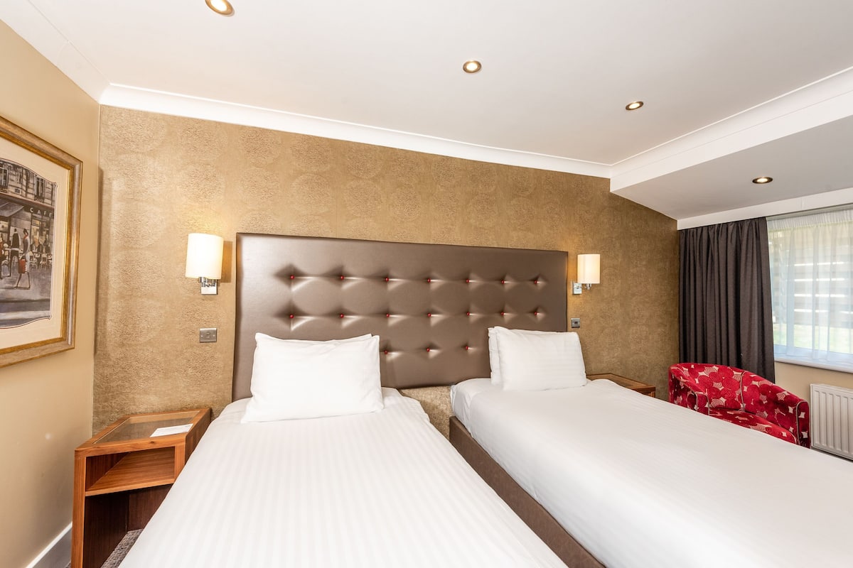 A hotel room with two beds, a large tufted headboard, wall-mounted lamps, and a framed picture on the wall.