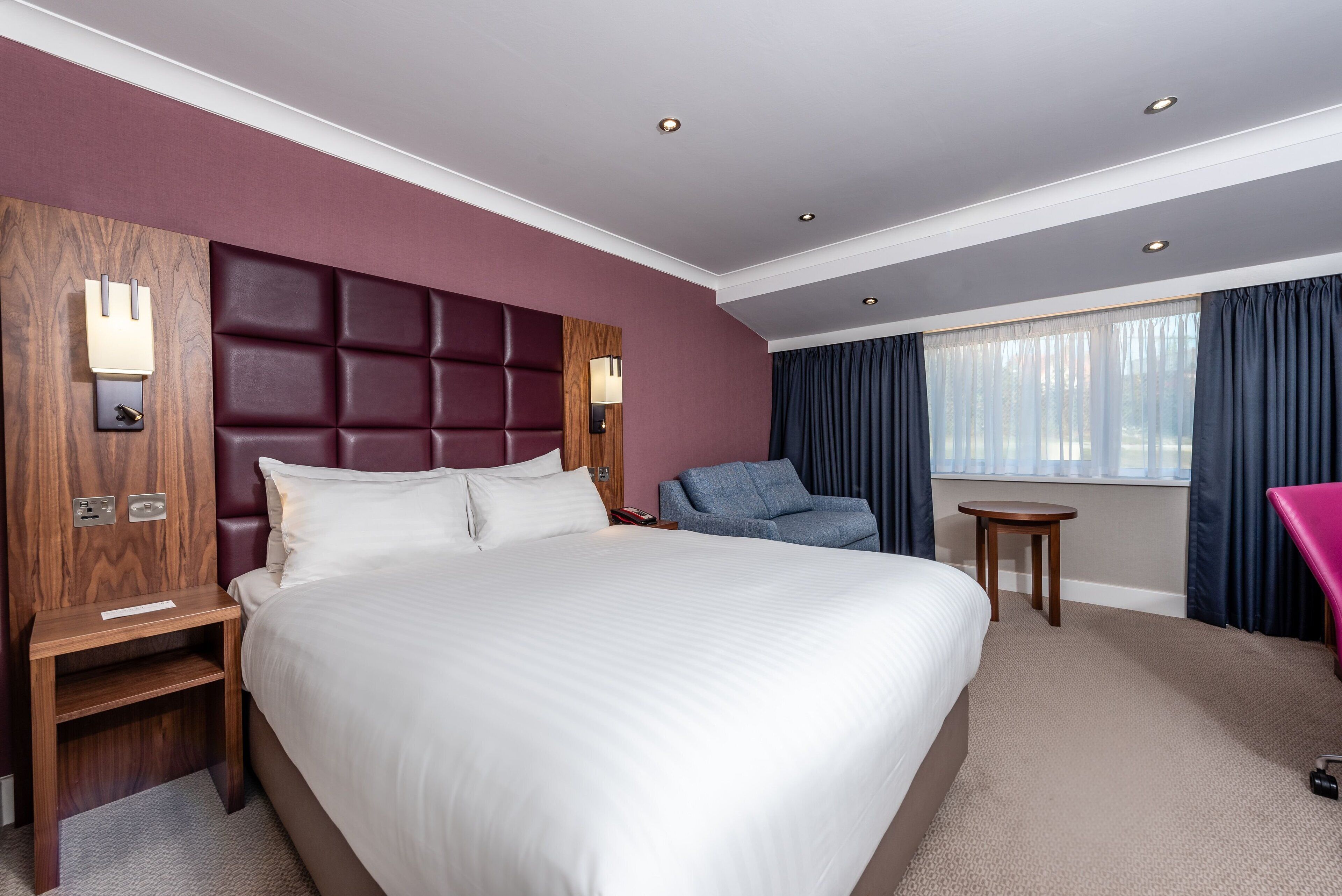 Habitación de hotel con una cama grande, una mesita de noche de madera, un sillón azul, una pequeña mesa y una ventana con cortinas.