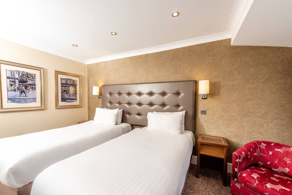 Een hotelkamer met twee bedden, een rode fauteuil en afbeeldingen in lijsten aan de muur.