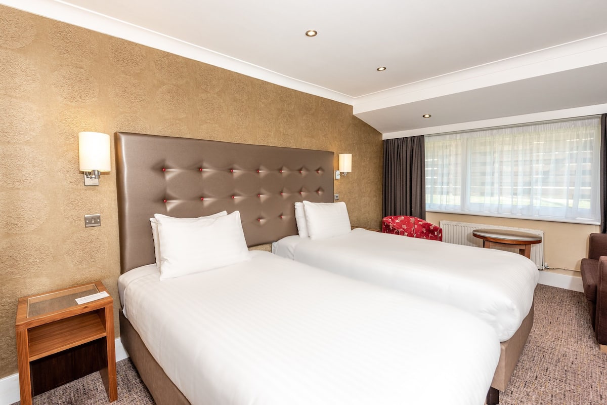 A hotel room with two beds, a headboard, bedside lamps, a small table, a chair, and a window with curtains.