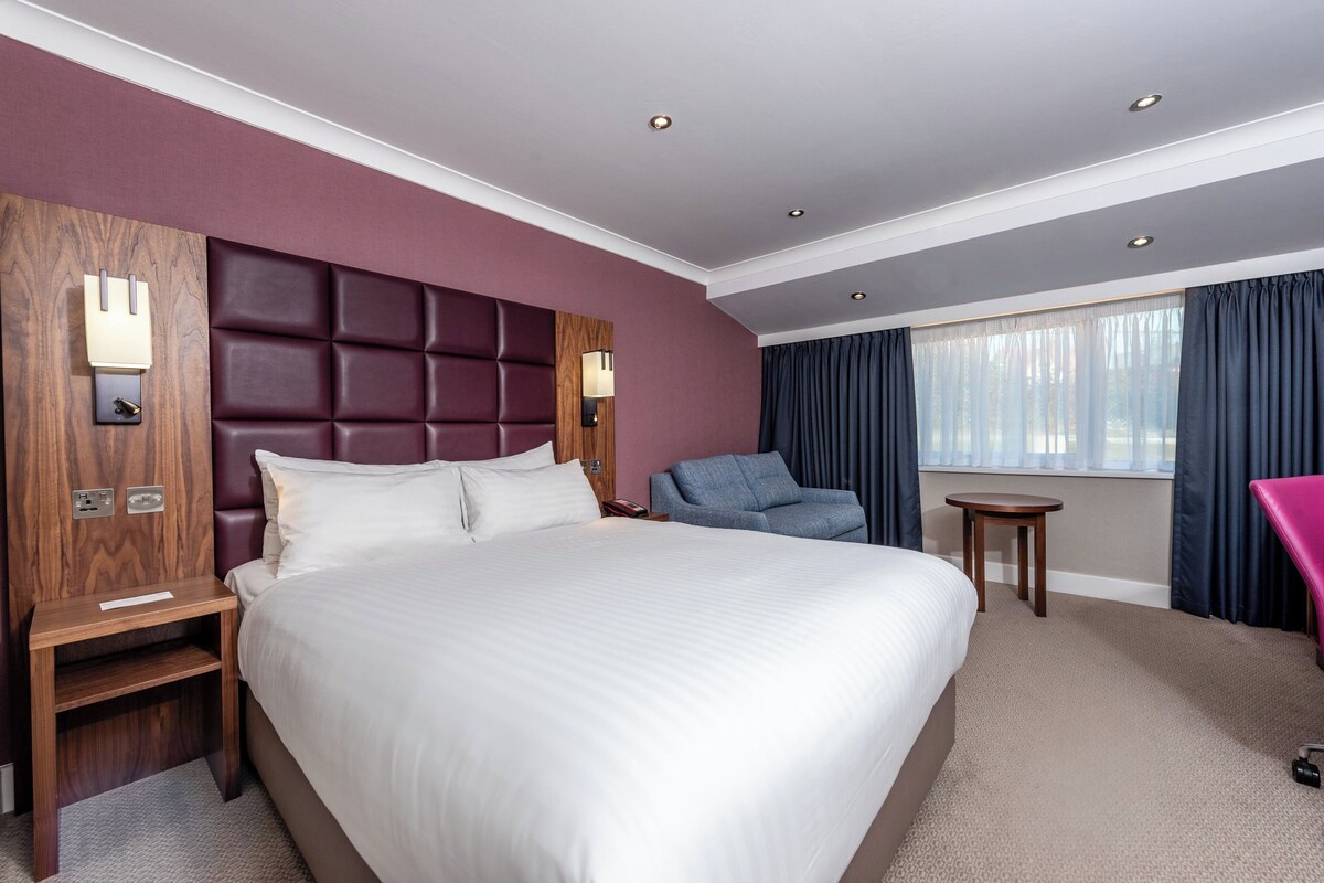 A hotel room with a large bed, a wooden nightstand, a blue armchair, a small table, and a window with curtains.