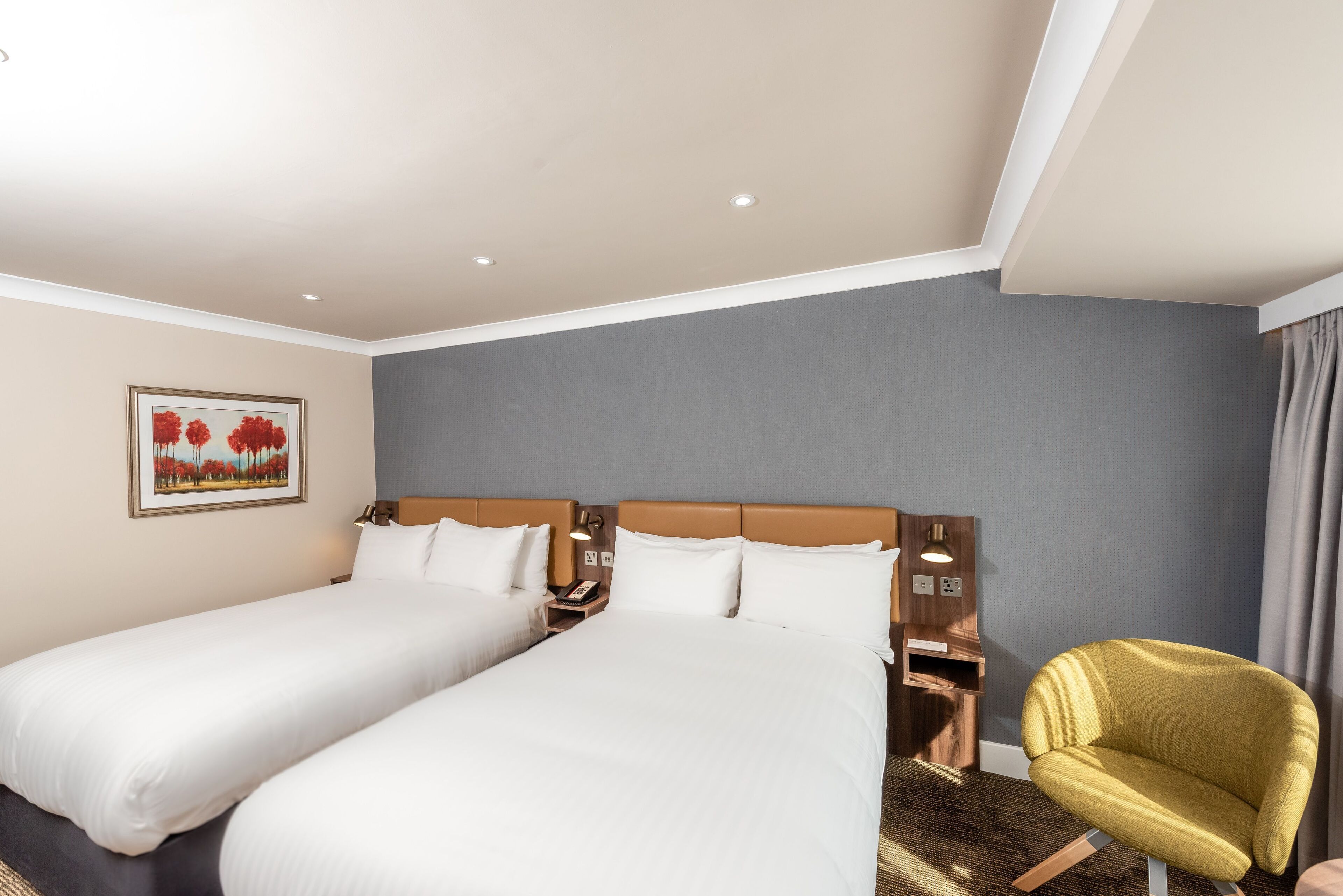Habitación de hotel con dos camas, una mesita de noche, una lámpara, una silla y un cuadro en la pared.