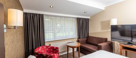 A hotel room with a bed, a red armchair, a brown sofa, a small round table, a TV, and a window with curtains.