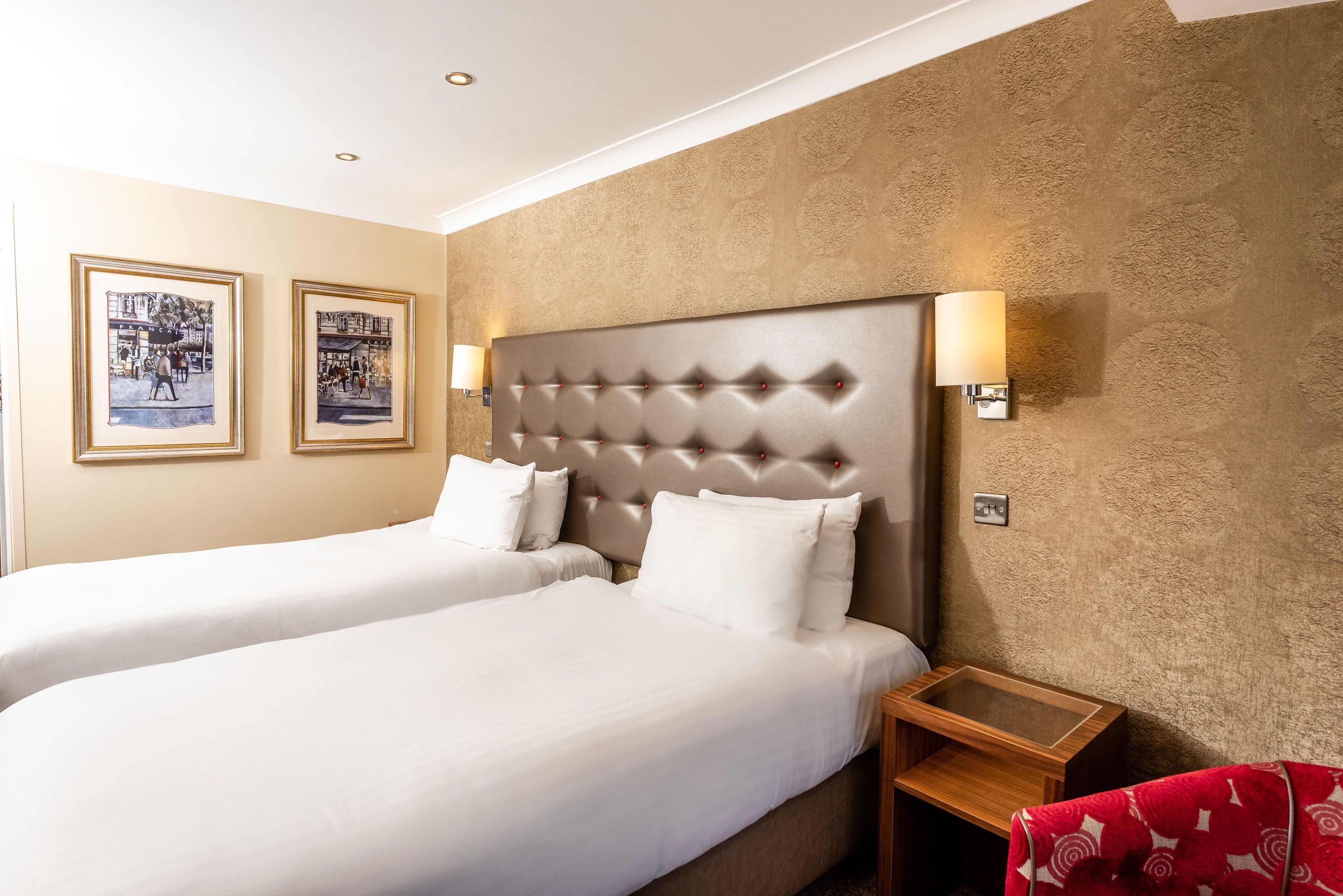 Una habitación de hotel con dos camas, cabeceros, lámparas de pared y cuadros enmarcados.