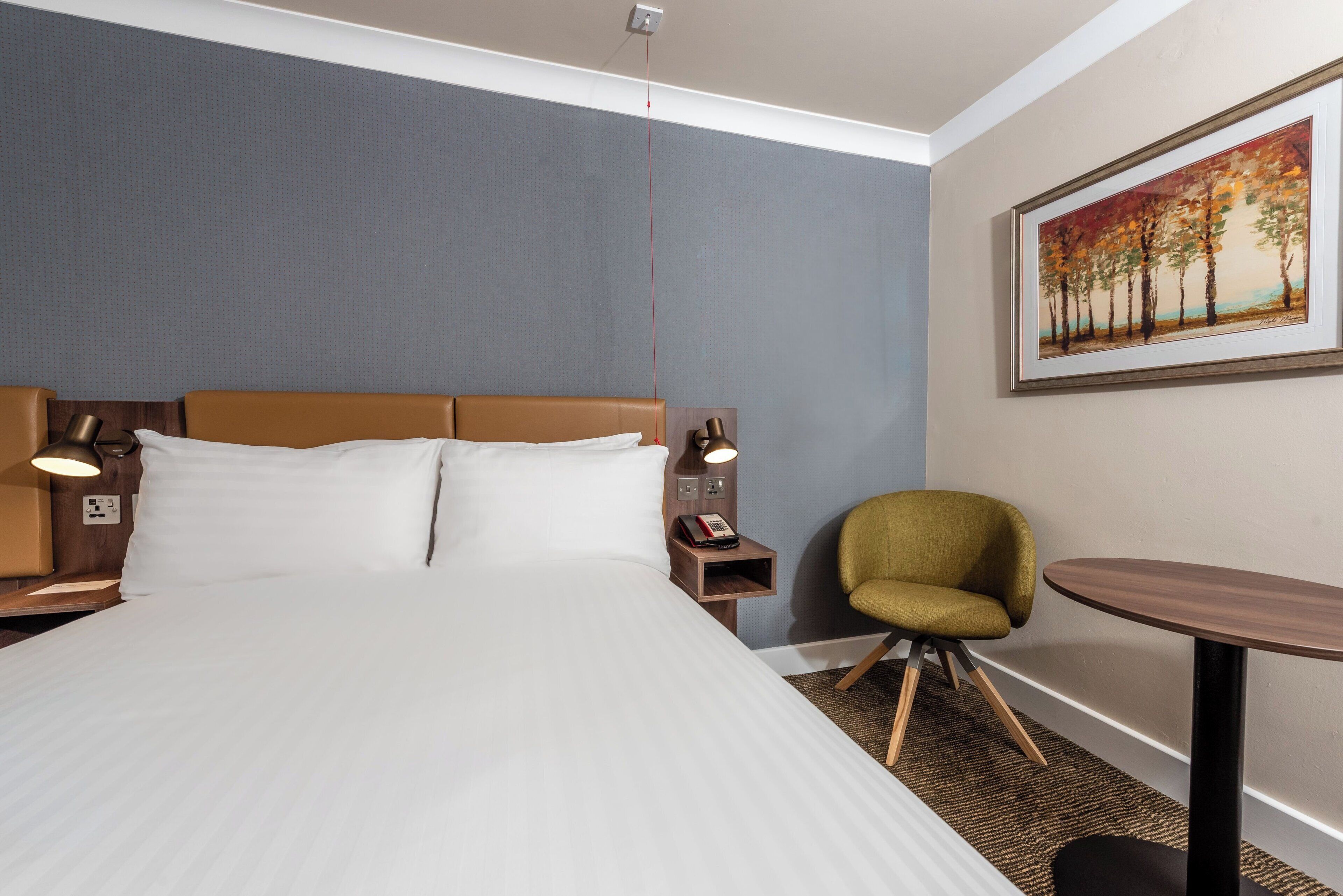 Una habitación de hotel con cama, lámparas de noche, una silla, una mesita y un cuadro en la pared.