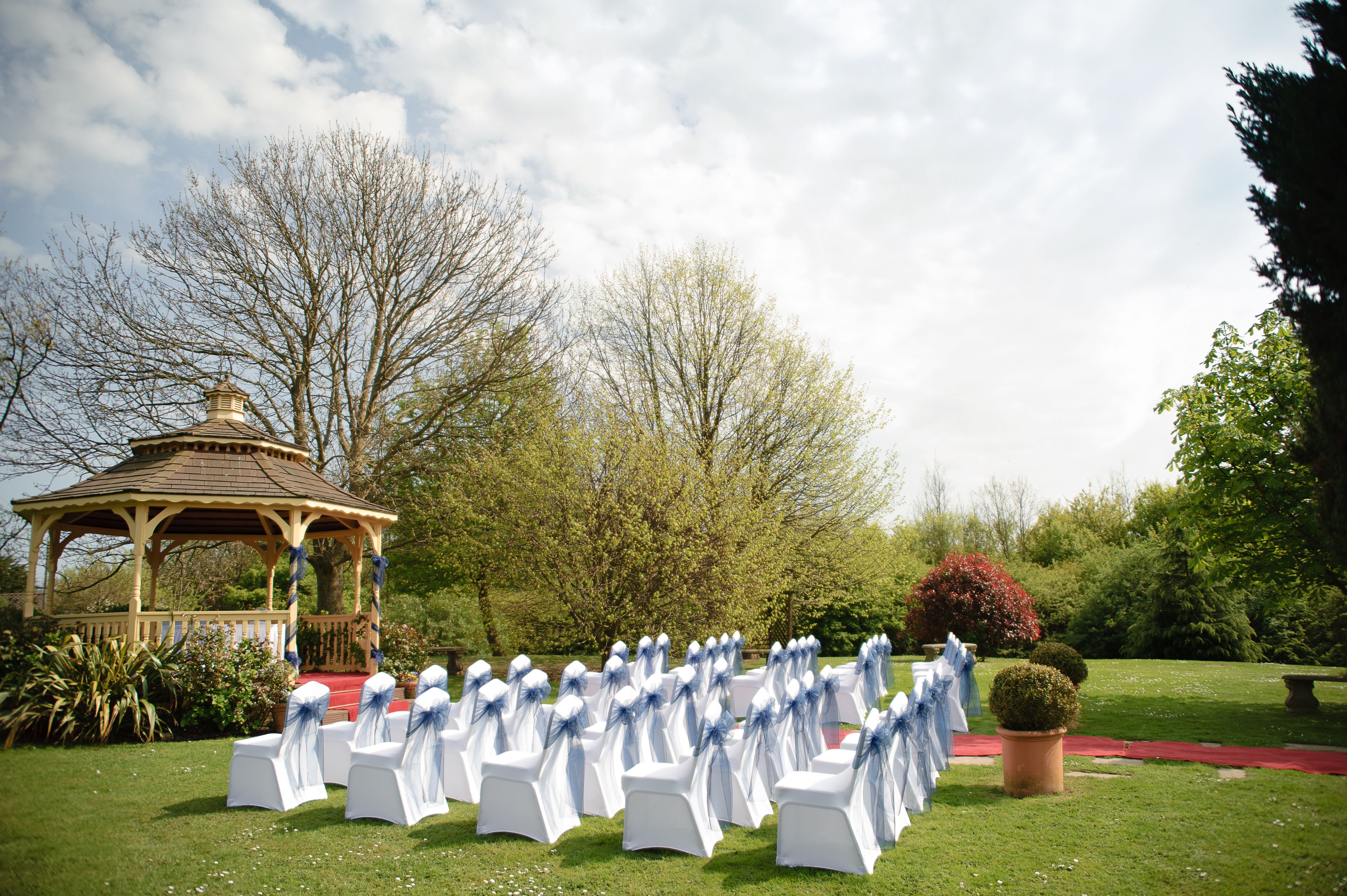 A gazebo with a red carpet leading to rows of white chairs with blue sashes, set in a garden with trees and a potted plant.