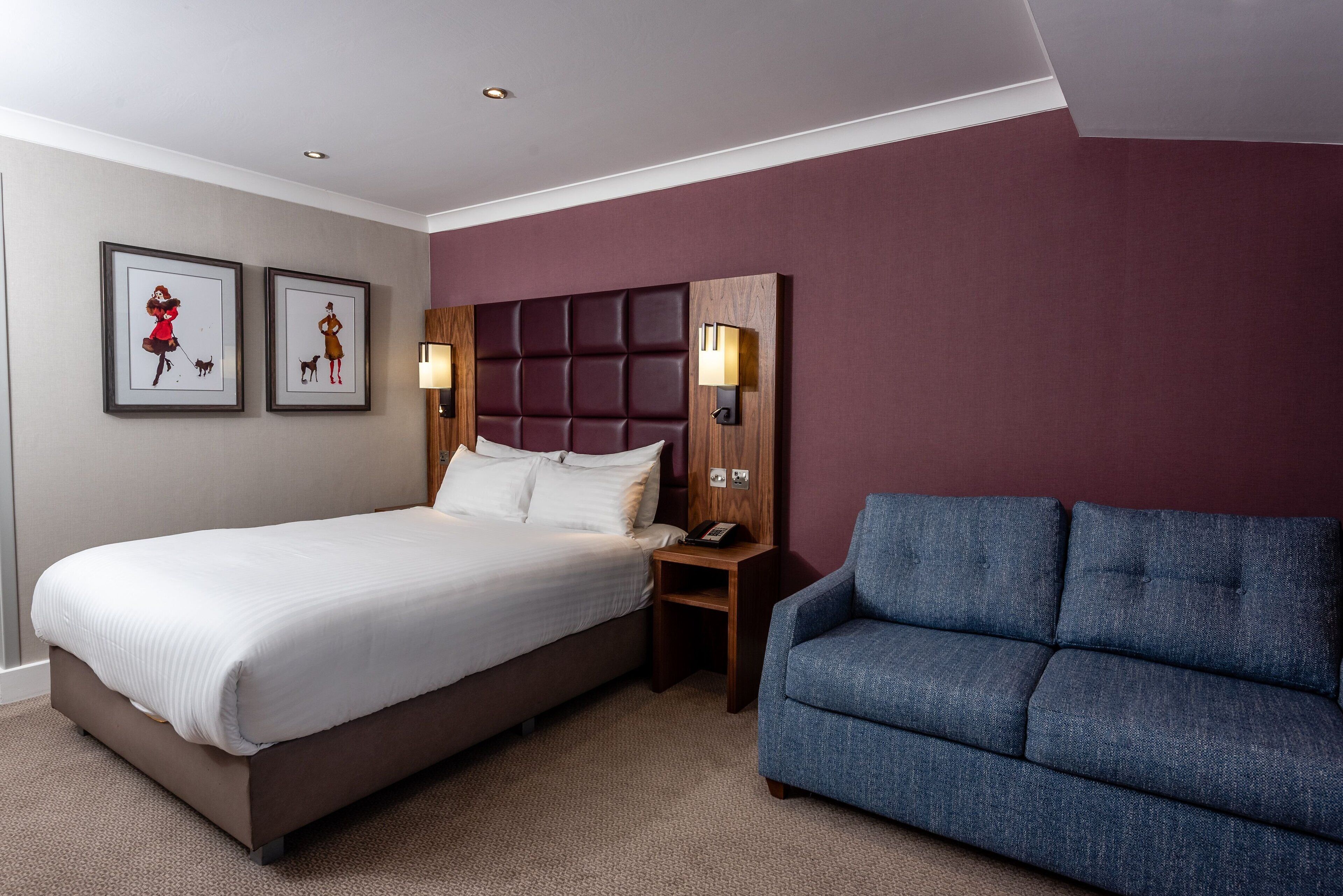 A hotel room with a bed, a blue sofa, and two framed pictures on the wall.