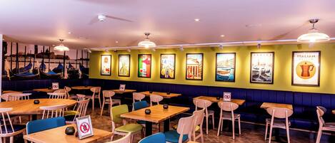 L’intérieur d’un restaurant présente de nombreuses tables et chaises, un mur orné de tableaux encadrés et un plafond doté d’un éclairage encastré.