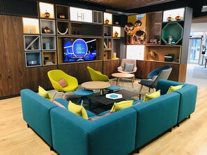 A modern lounge area with teal sofas, colorful chairs, and a central coffee table. A television is mounted on the wall displaying a quiz show. - Holiday Inn Dover by IHG (Dover)