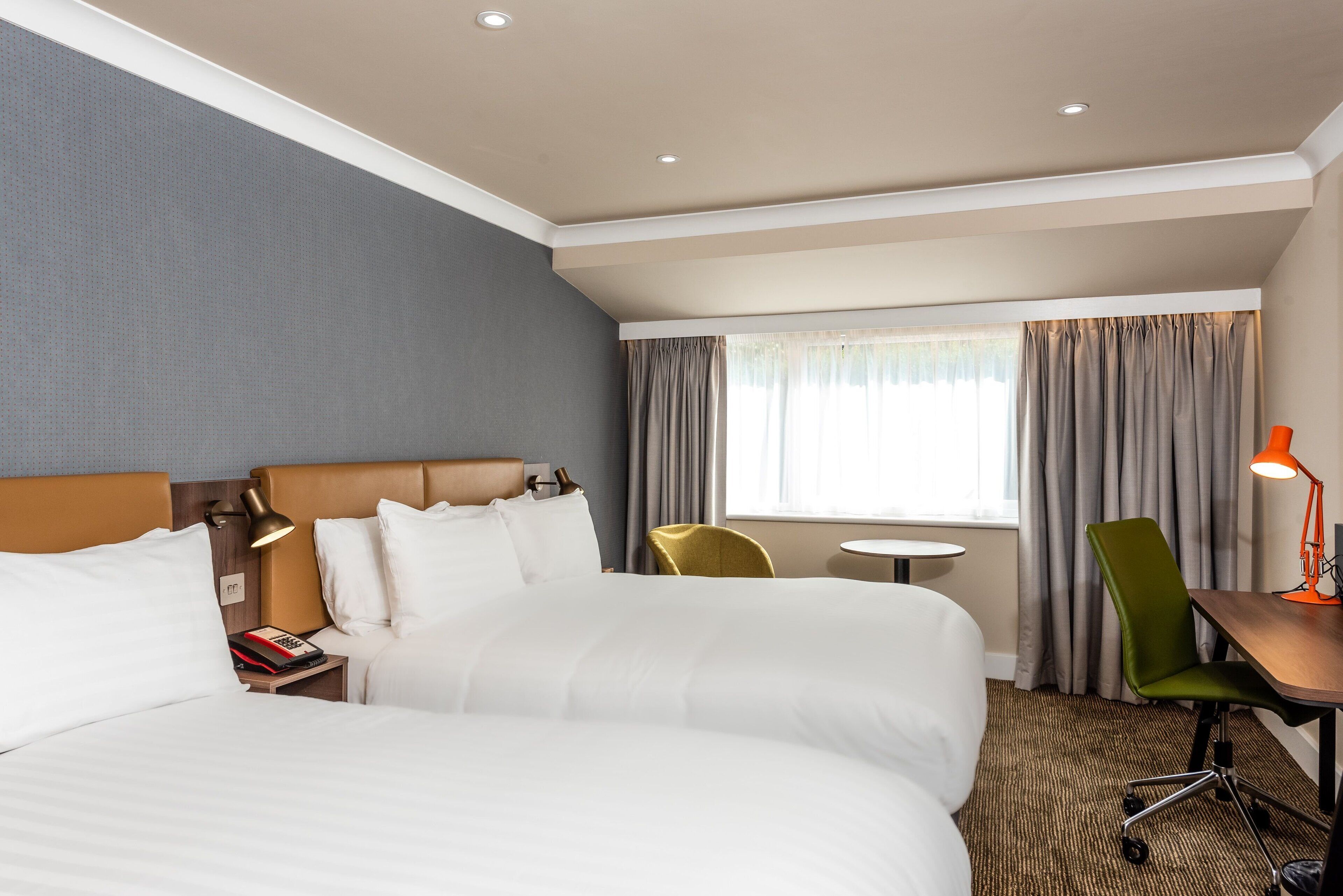 A hotel room with a large bed, a desk with a lamp, a chair, and a small table.