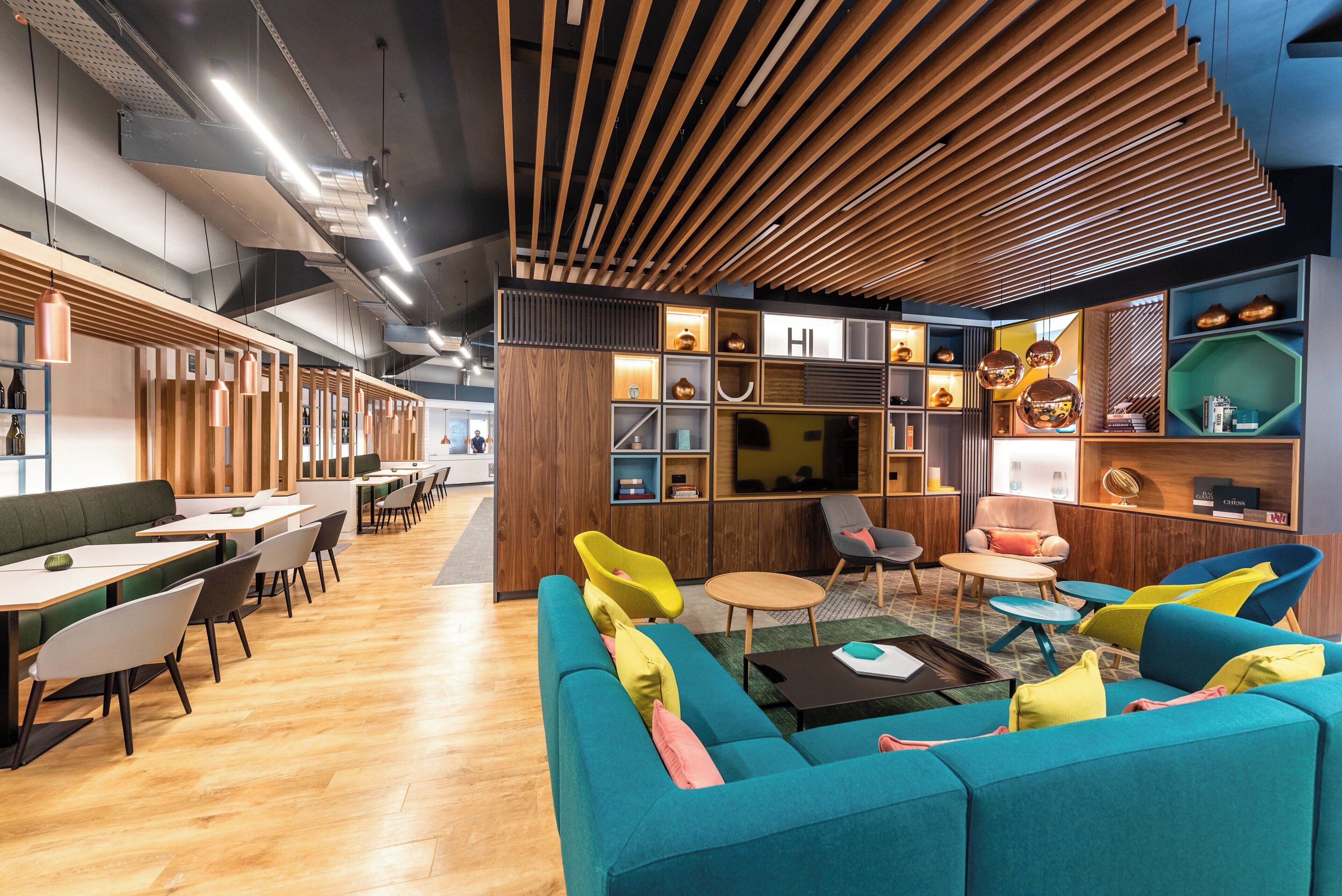 Un interior moderno con listones de madera en el techo, una zona de estar central con cojines de colores y un televisor fijado a la pared.