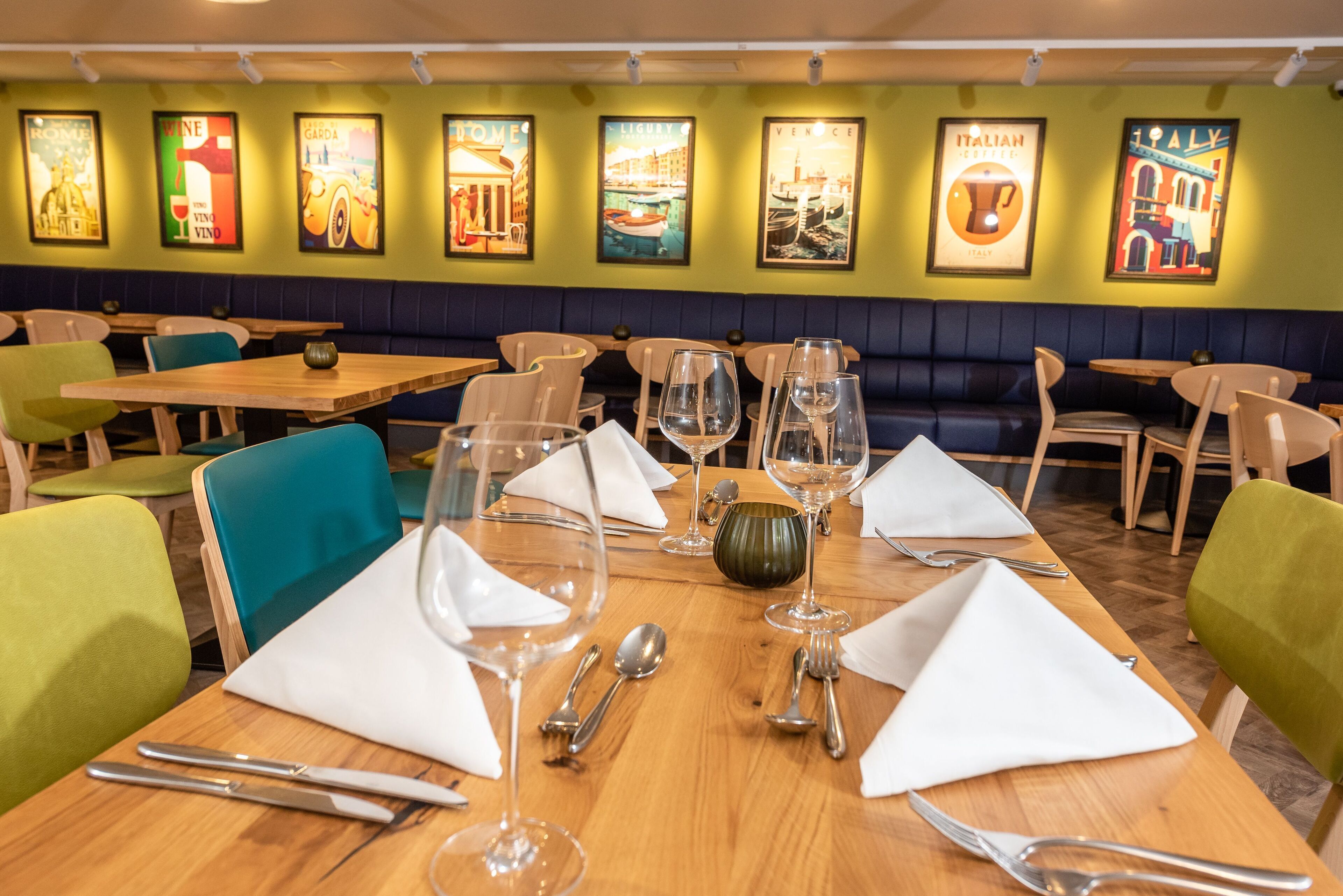 Un restaurante con mesas de madera preparadas para comer, sillas verdes y azules, y cuadros en la pared que muestran lugares de Italia.