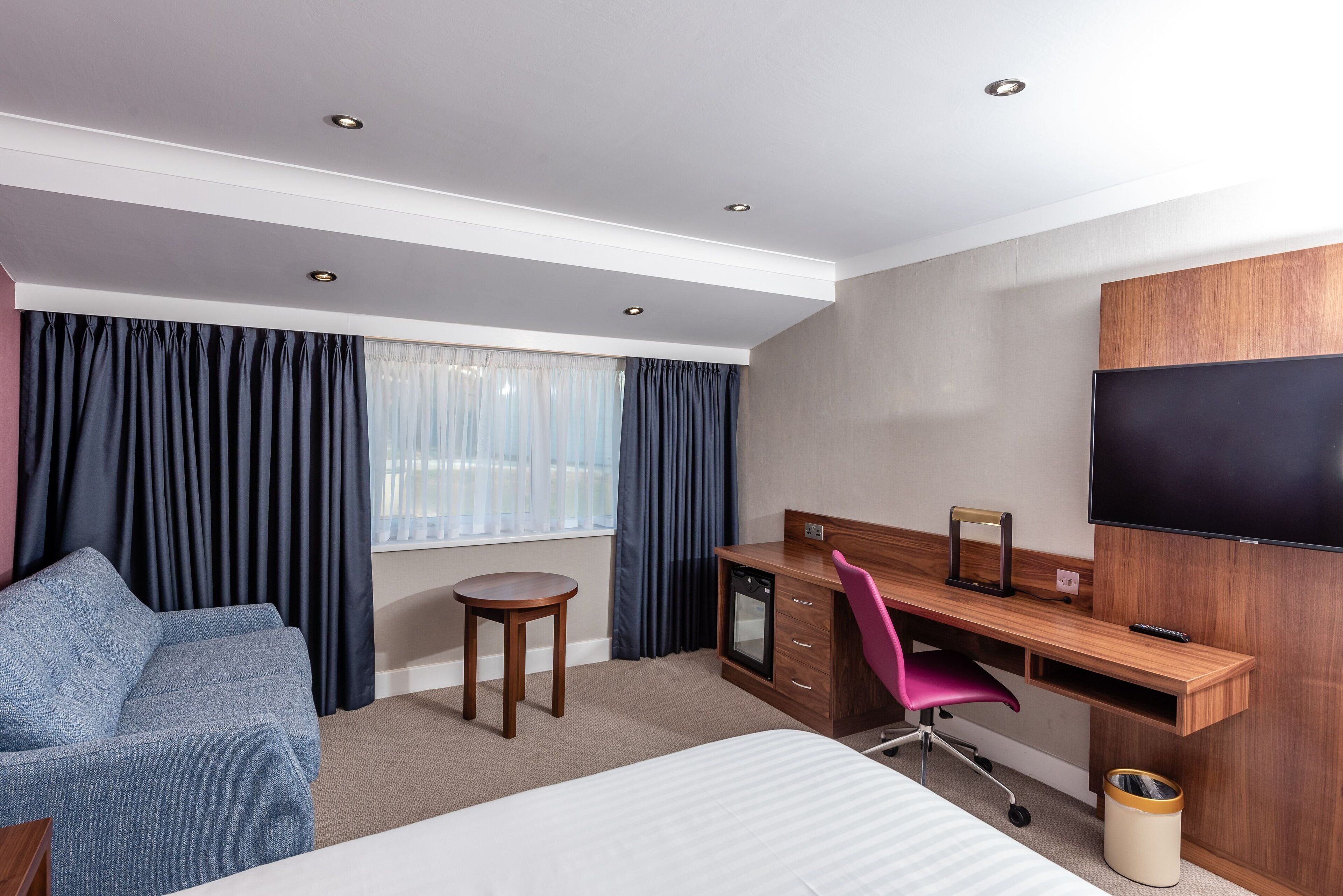 Habitación de hotel con una cama, un escritorio con computadora, una silla, un televisor y una ventana con cortinas.