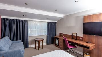 Habitación de hotel con una cama, un escritorio con computadora, una silla, un televisor y una ventana con cortinas.