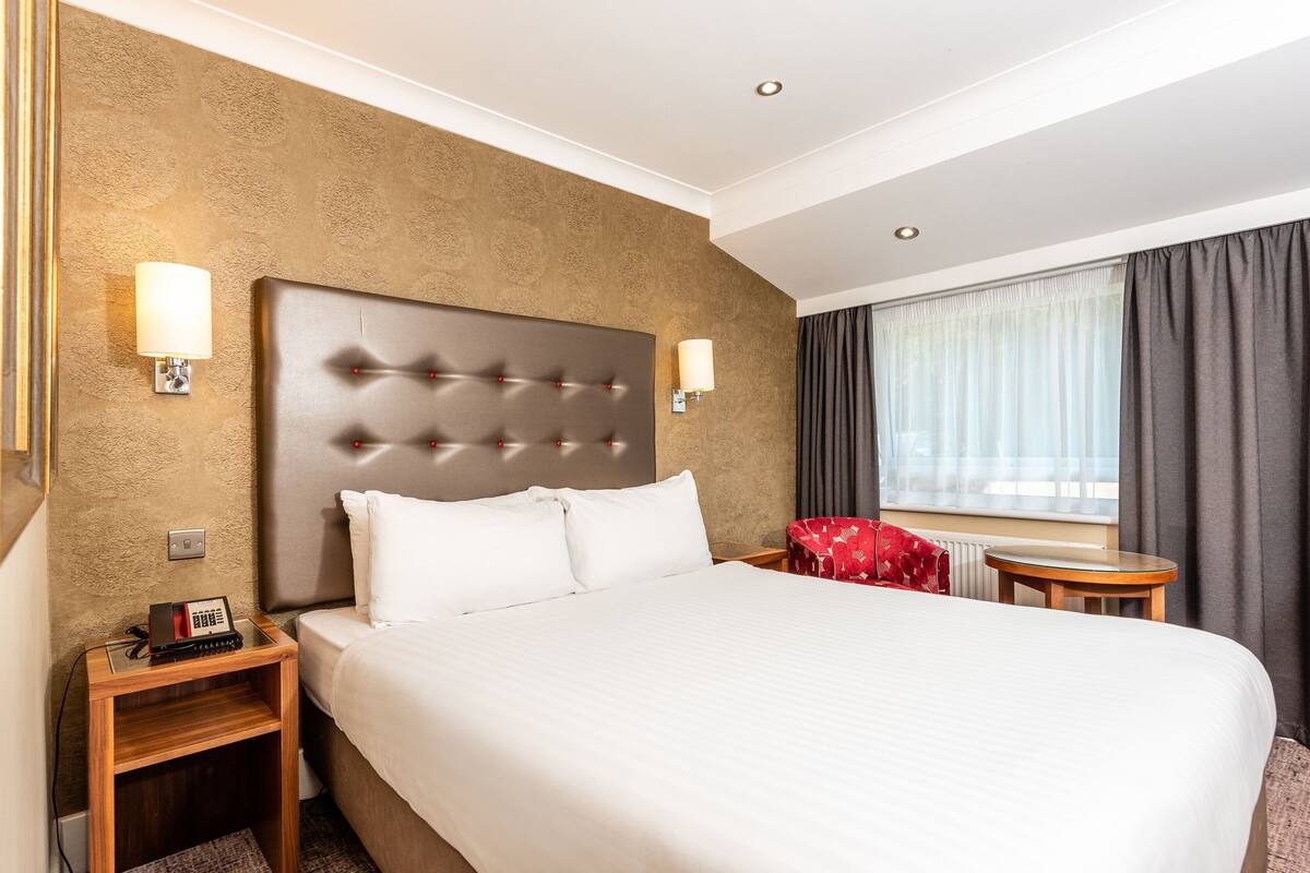 Een hotelkamer met een groot bed, twee nachtkastjes, een rode stoel en een raam met gordijnen.