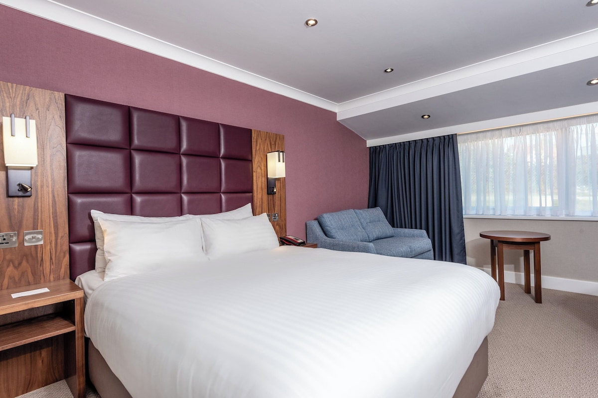 Habitación de hotel con una cama grande, un cabecero de madera, una zona de estar con una silla, una mesita y una ventana con cortinas.