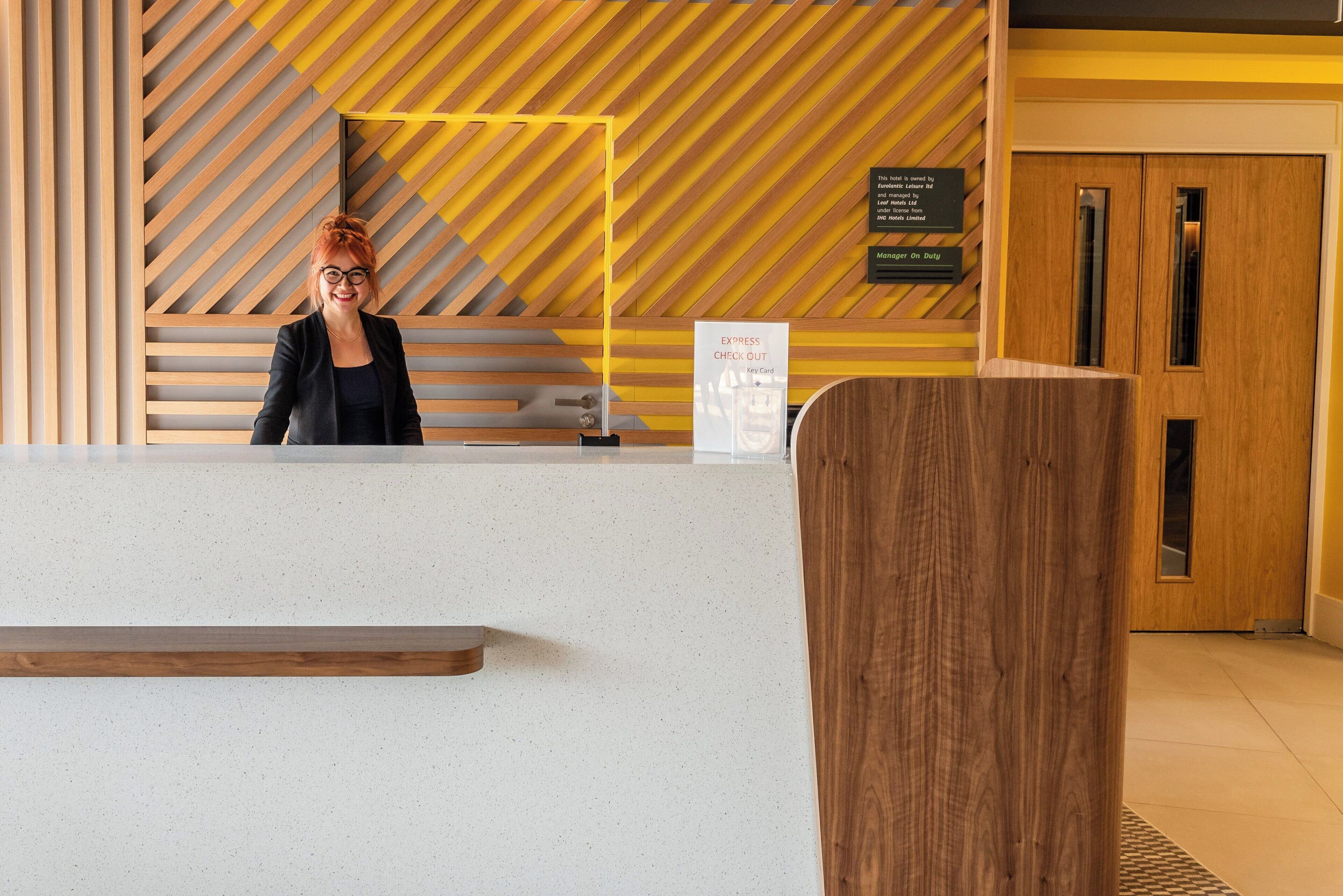 A reception desk with a modern design, a sign indicating check-out services, and a wooden door in the background.