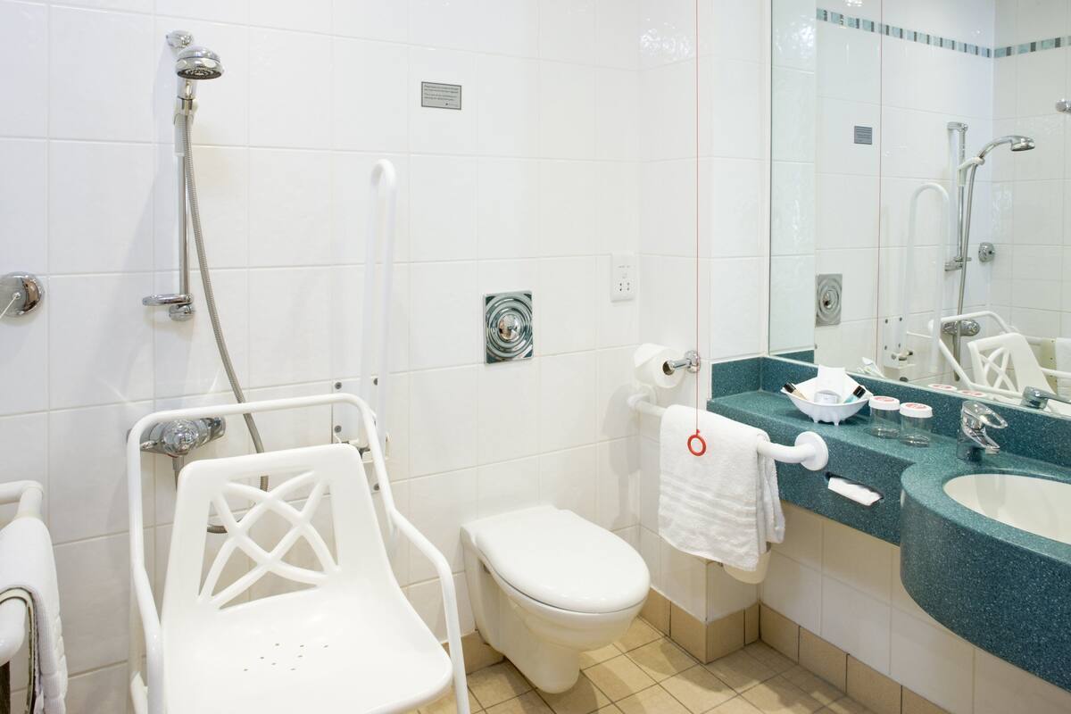Een moderne badkamer met een wit toilet, een douche met een handdouche, een witte stoel, een wastafel met een spiegel en een handdoekrek.