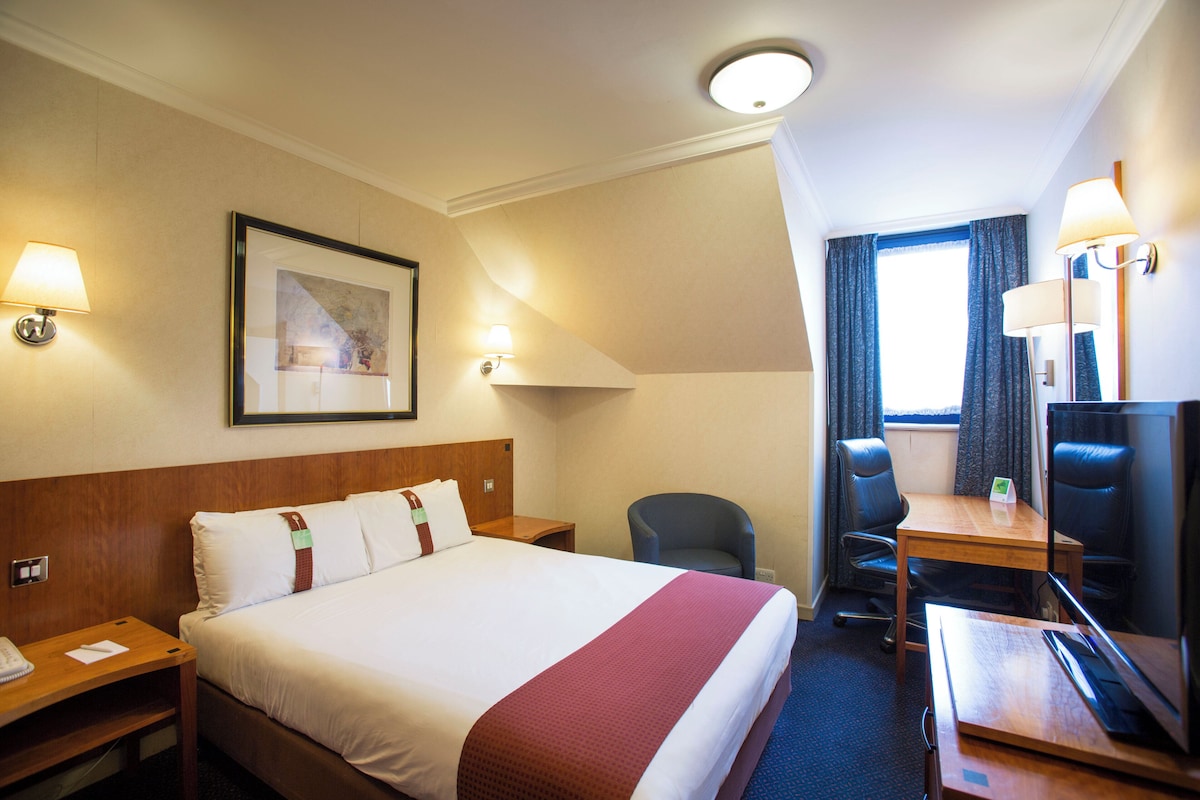 A hotel room with a bed, bedside tables, a desk, a chair, a television, and a window with curtains.