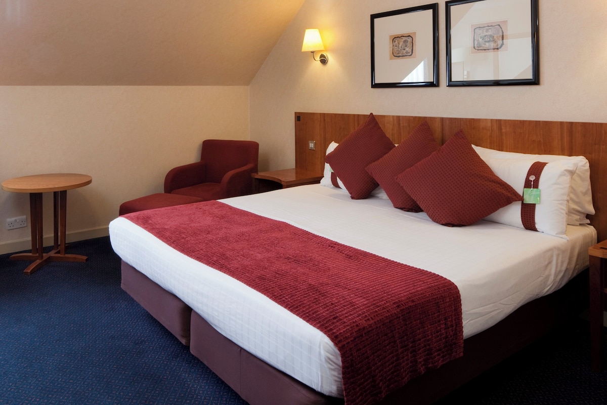A hotel room with a bed, a chair, a small table, and two framed pictures on the wall.