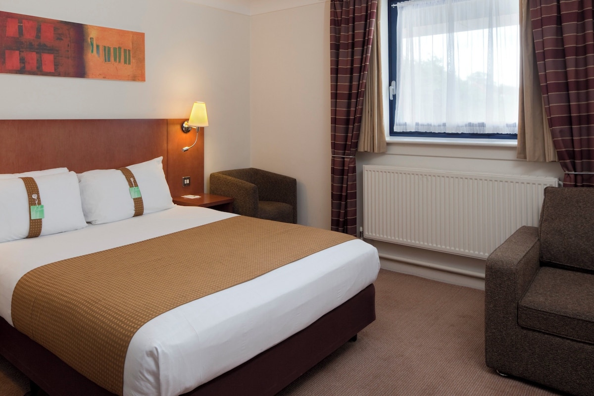 A hotel room with a large bed, a nightstand, a chair, a window with curtains, and a wall-mounted lamp.