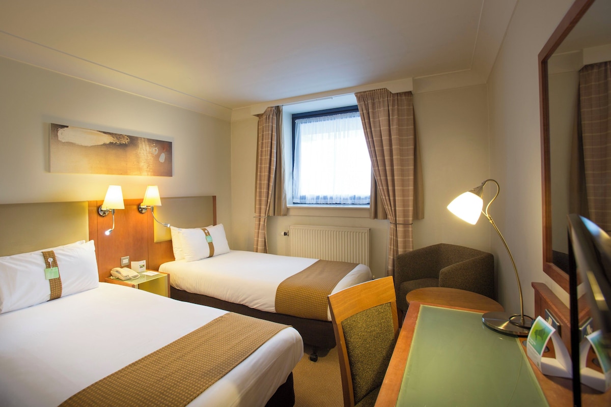 A hotel room with two beds, a desk, a chair, a lamp, a window with curtains, and a painting on the wall.