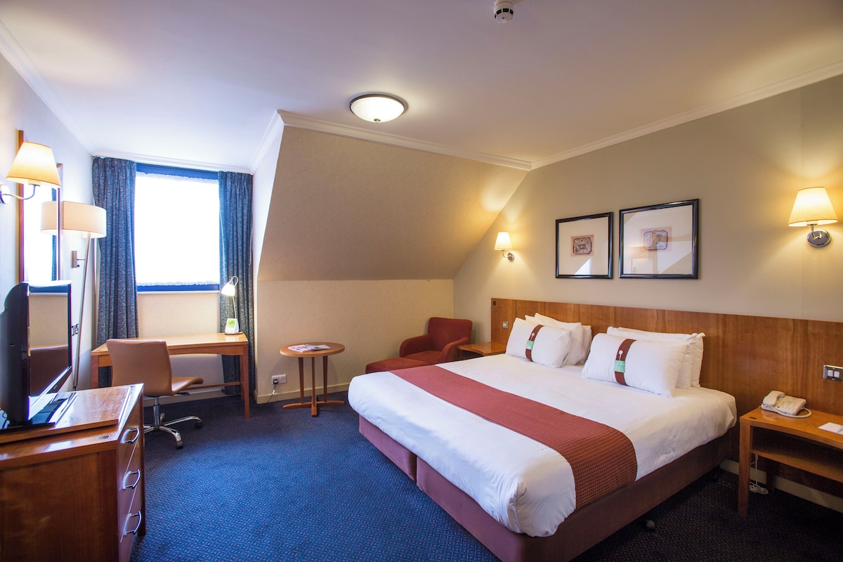 A hotel room with a large bed, a desk, a chair, a small table, and a telephone.