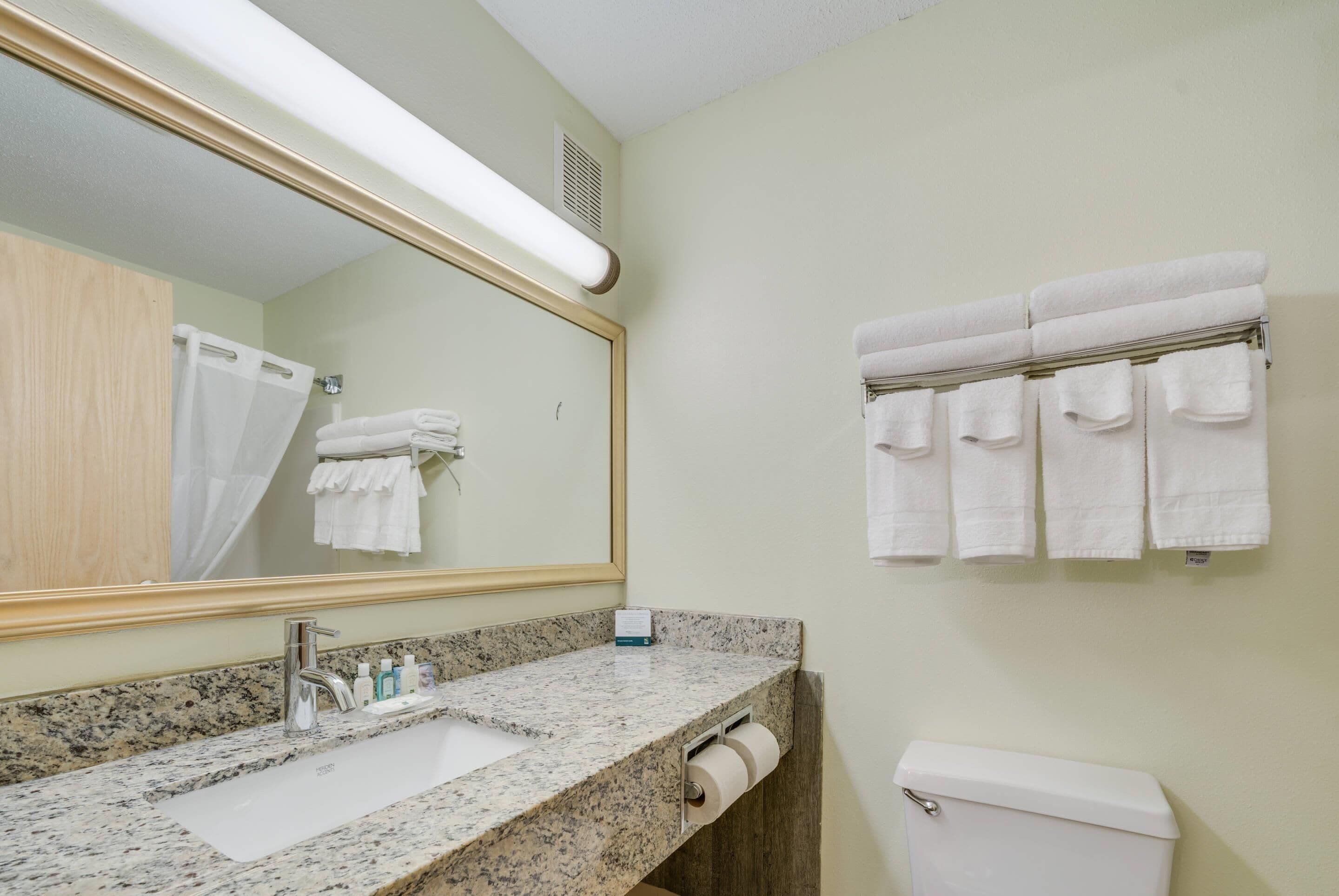 A bathroom with a granite countertop sink, a large mirror, a toilet, and a towel rack with multiple towels.