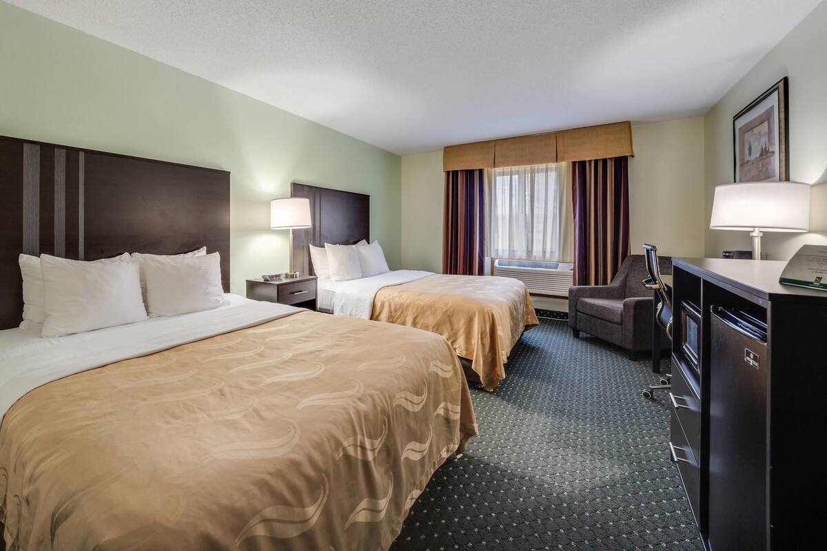 A hotel room with two beds, a desk, a chair, a refrigerator, and a window with curtains.