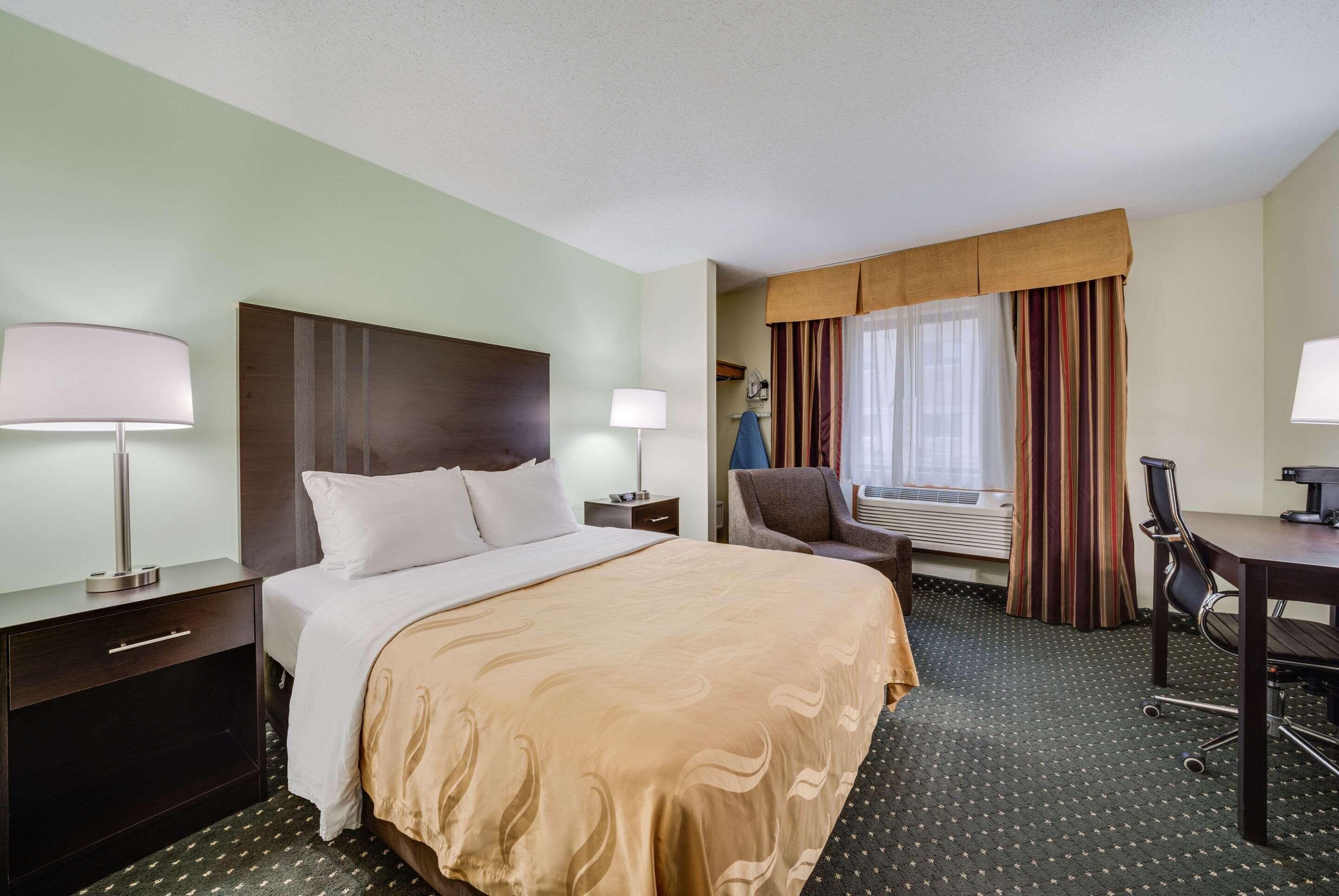A hotel room with a large bed, two bedside tables with lamps, a desk with a chair, a window with curtains, and a carpeted floor.