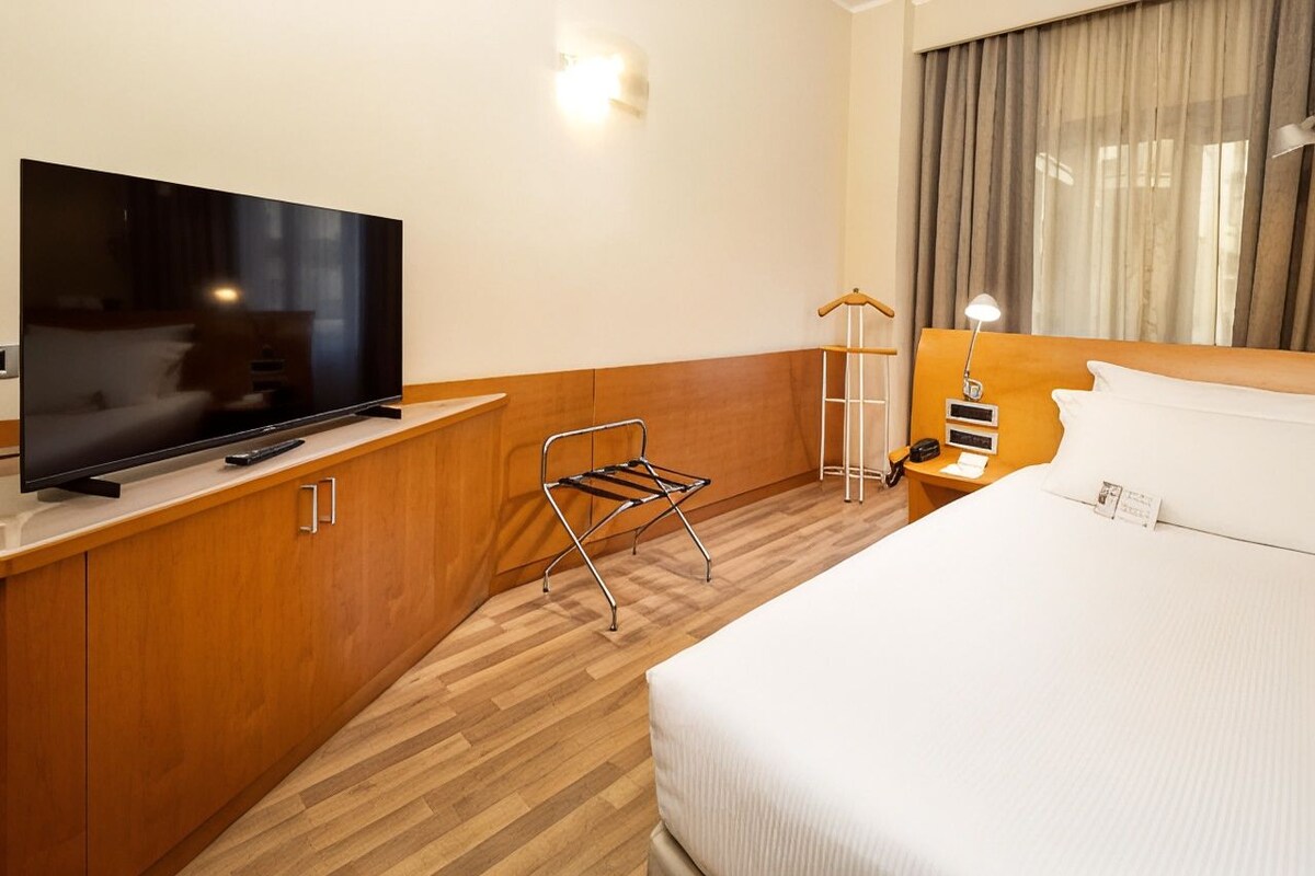 Une chambre d’hôtel avec un téléviseur à écran plat posé sur un meuble en bois, un lit avec du linge de maison blanc, une table de chevet avec une lampe et une fenêtre avec des rideaux.