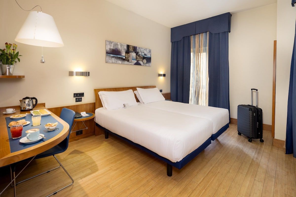 A hotel room with a bed, a desk with breakfast, a lamp, a painting, and a suitcase.