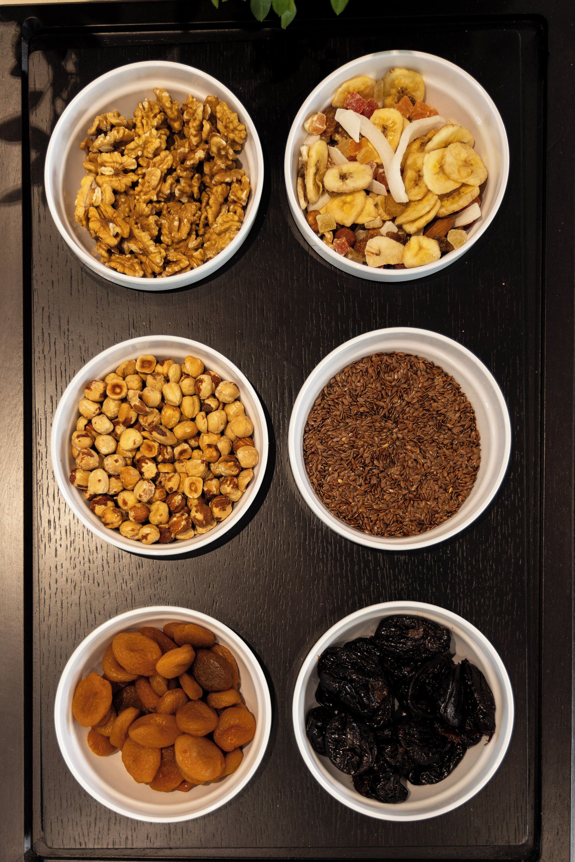 Four bowls of different nuts and dried fruits on a dark surface.