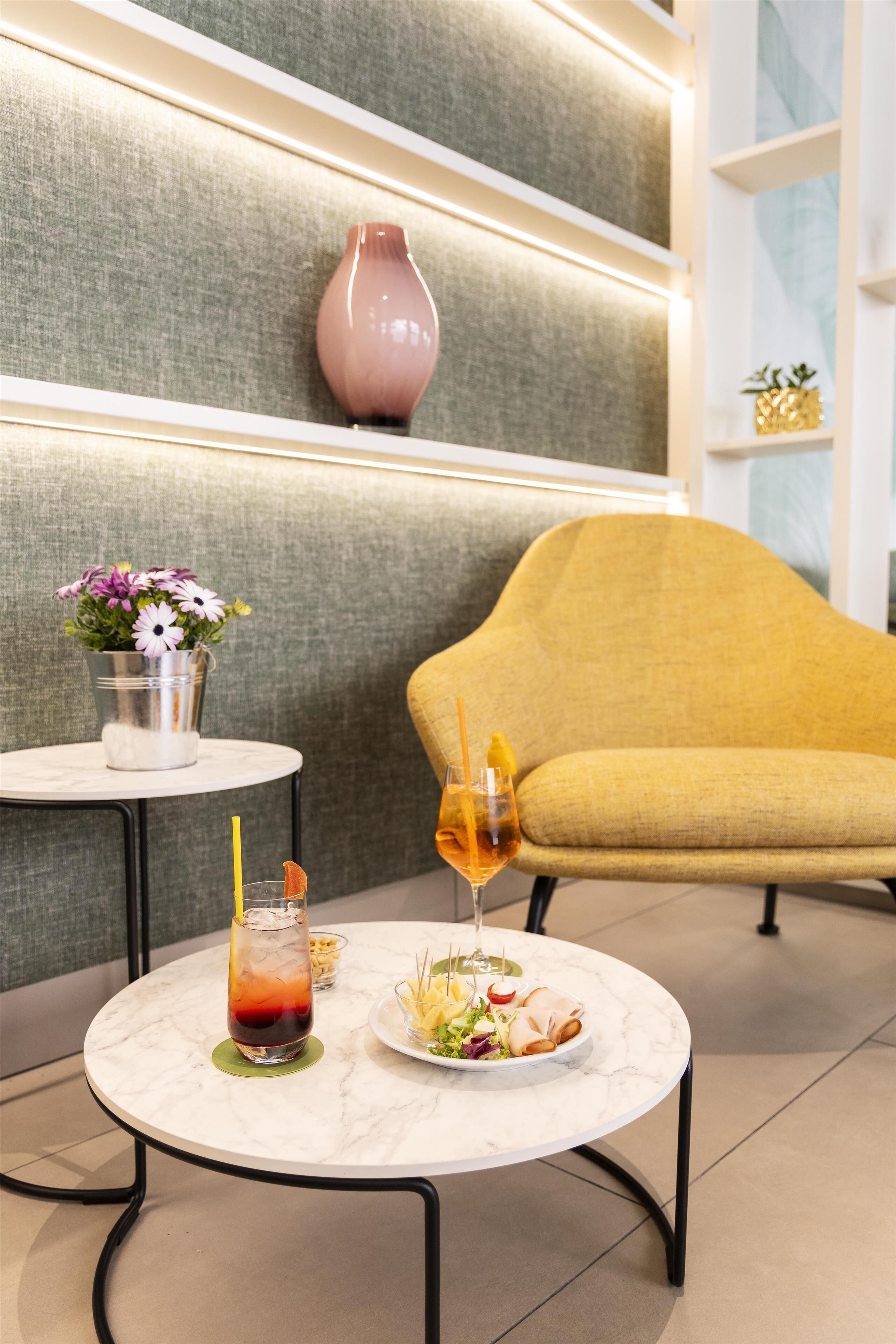 Un tavolo rotondo in marmo con bevande, un piatto di cibo, un vaso con fiori e una poltrona gialla in un ambiente moderno.